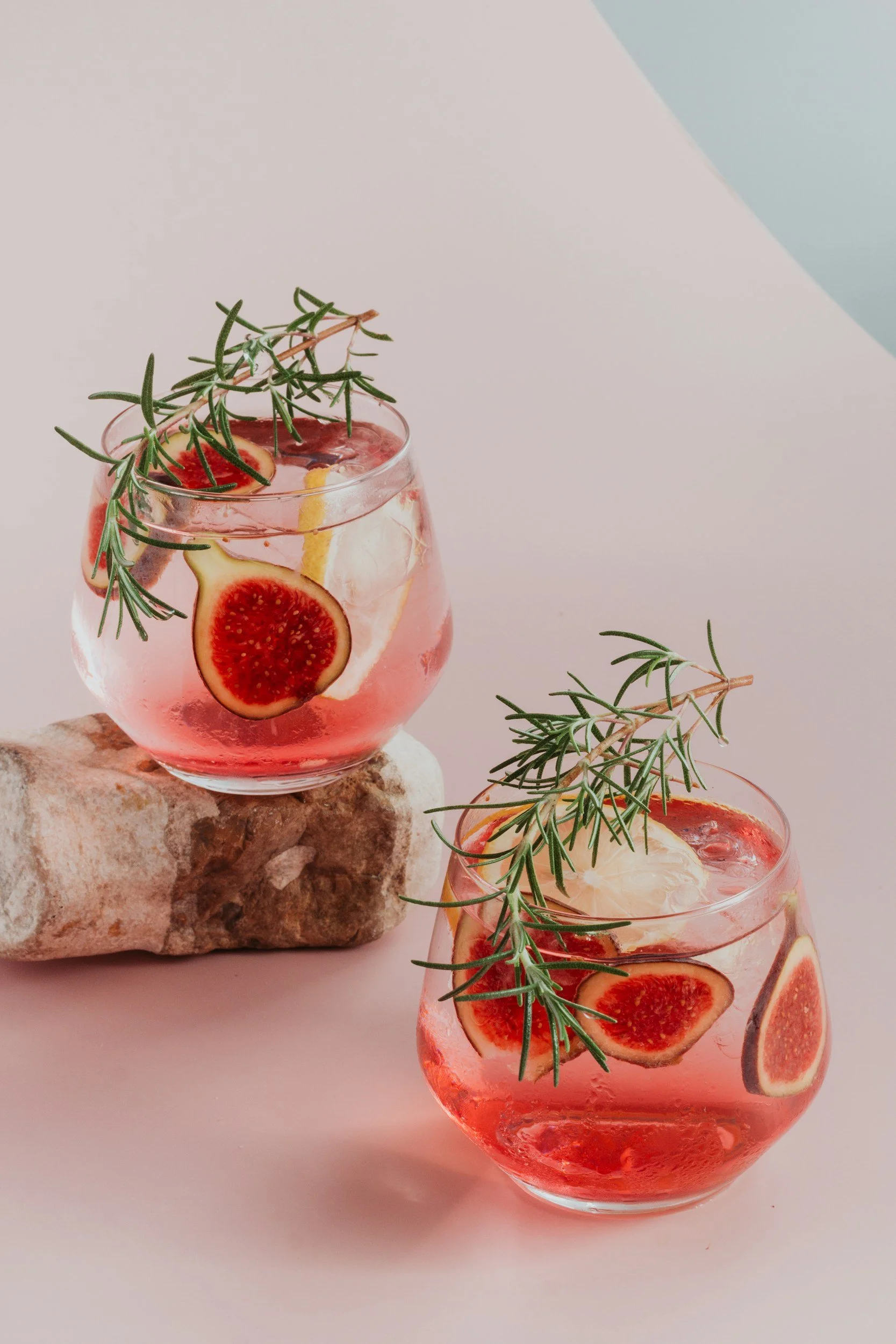 Two glasses of pink drink with fig slices, lemon slices, ice, and sprigs of rosemary, one glass is on a rock, against a pastel background.