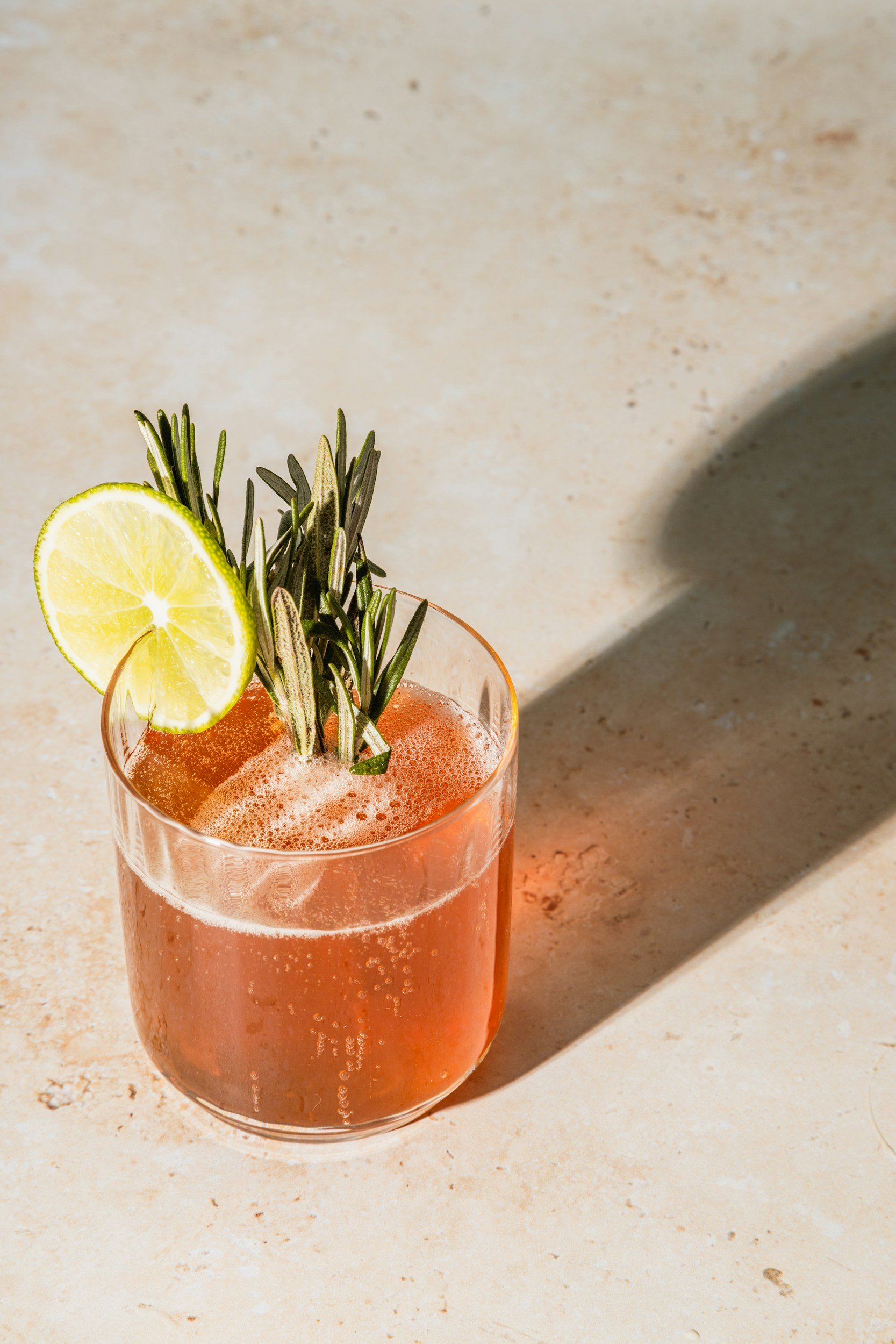 A glass of pink cocktail with foam, garnished with a lemon slice and sprigs of rosemary on a beige surface.