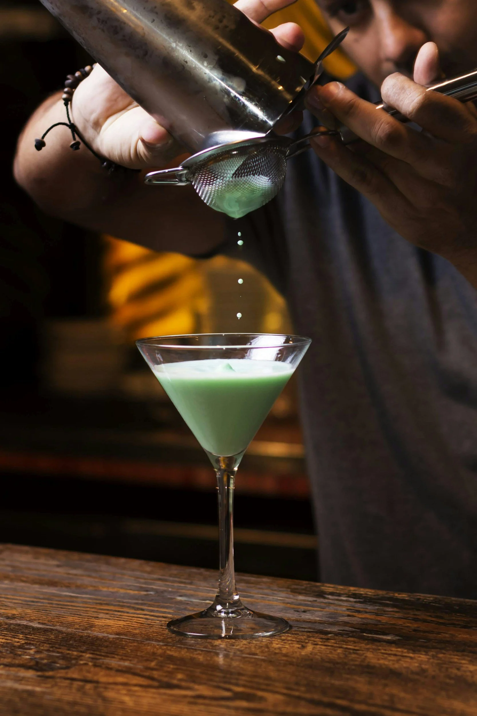 A bartender pours a green cocktail through a fine mesh strainer into a martini glass on a wooden bar counter.