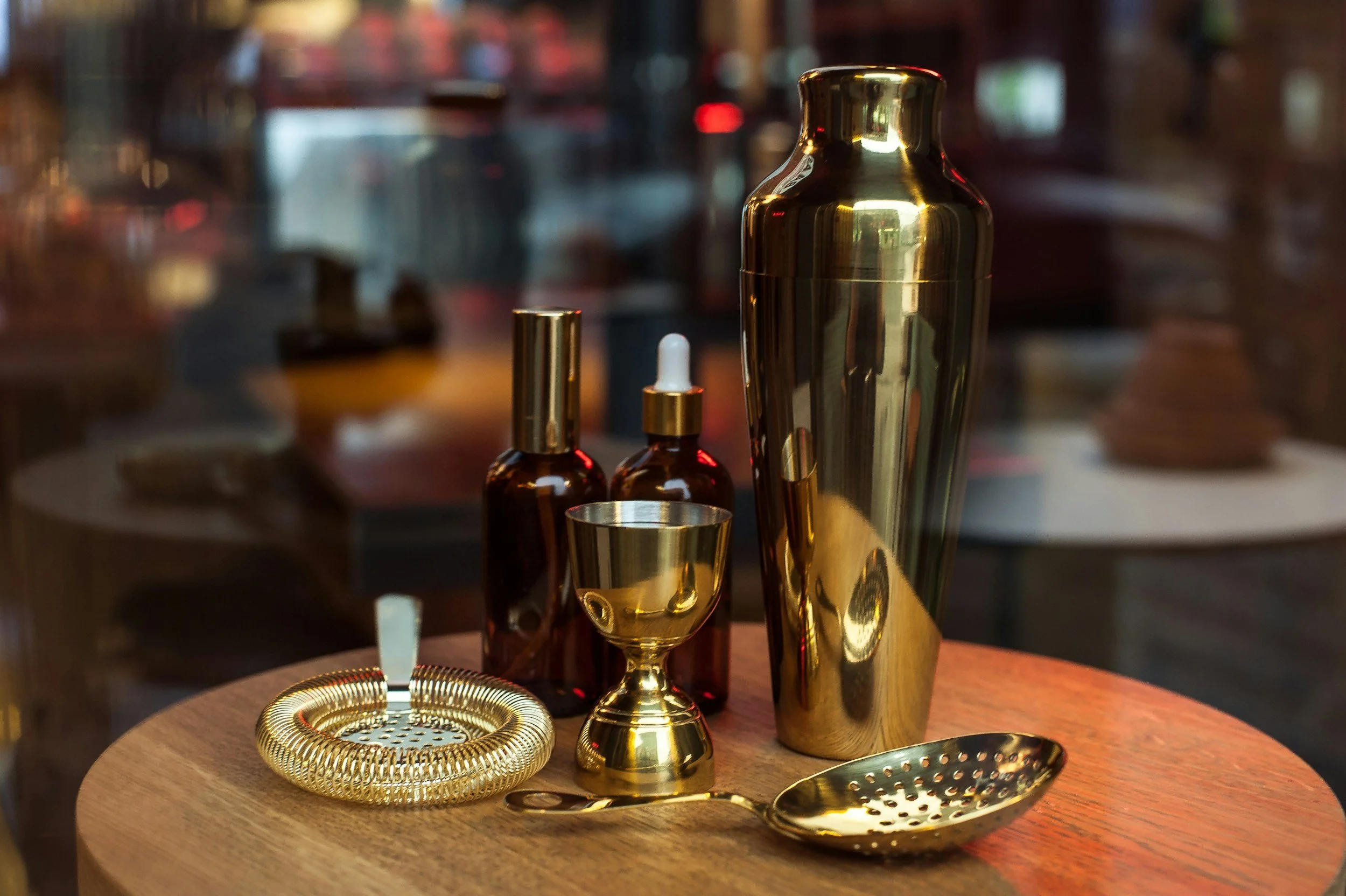 Various gold and brown cocktail-making tools and bottles on a wooden table, including a tall gold cocktail shaker, a small gold drinking cup, a gold strainer, a dropper bottle, a spray bottle, and a gold spoon.