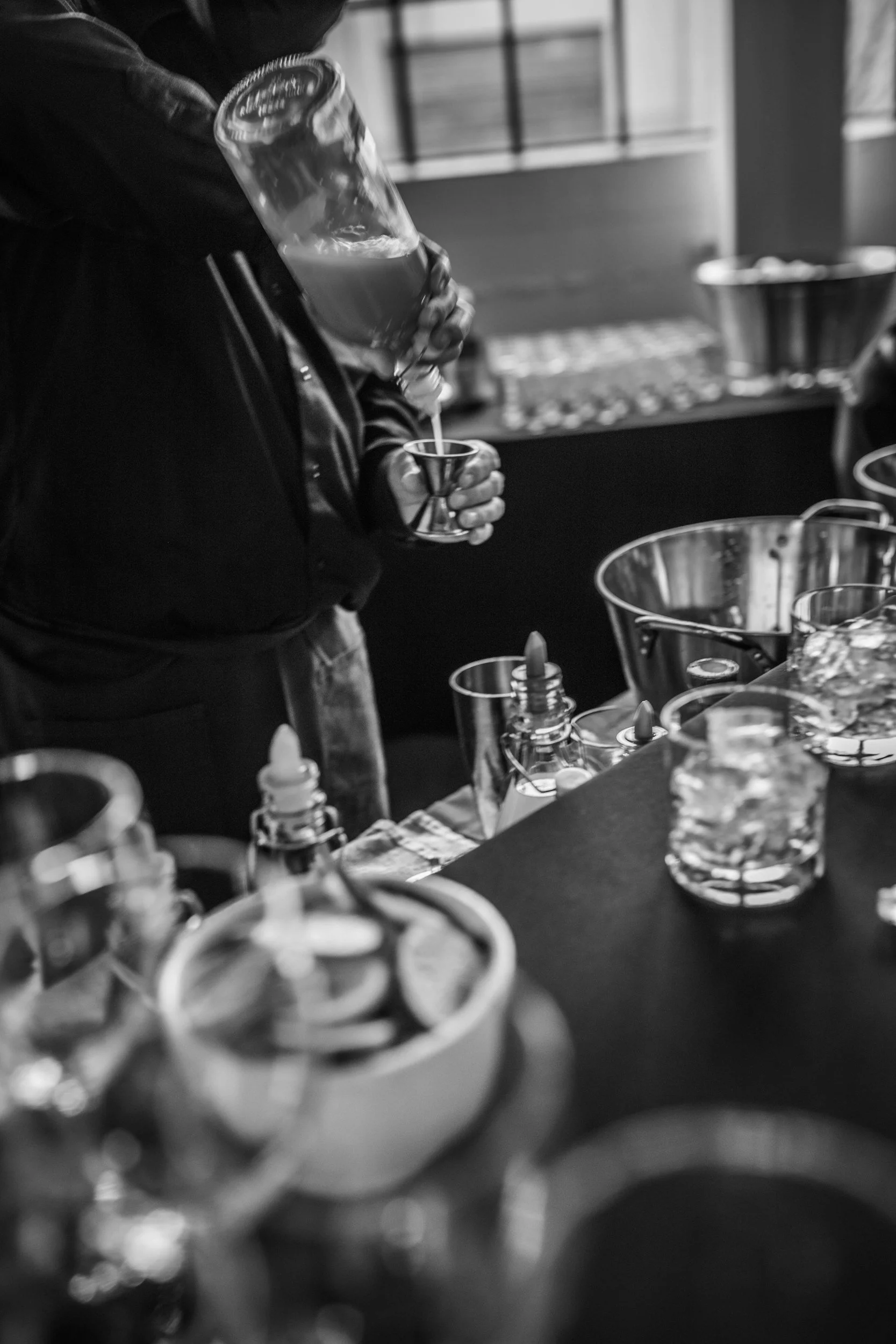 A person pouring liquid from a large bottle into a small glass, surrounded by various bottles, glasses, and bar tools on a table.