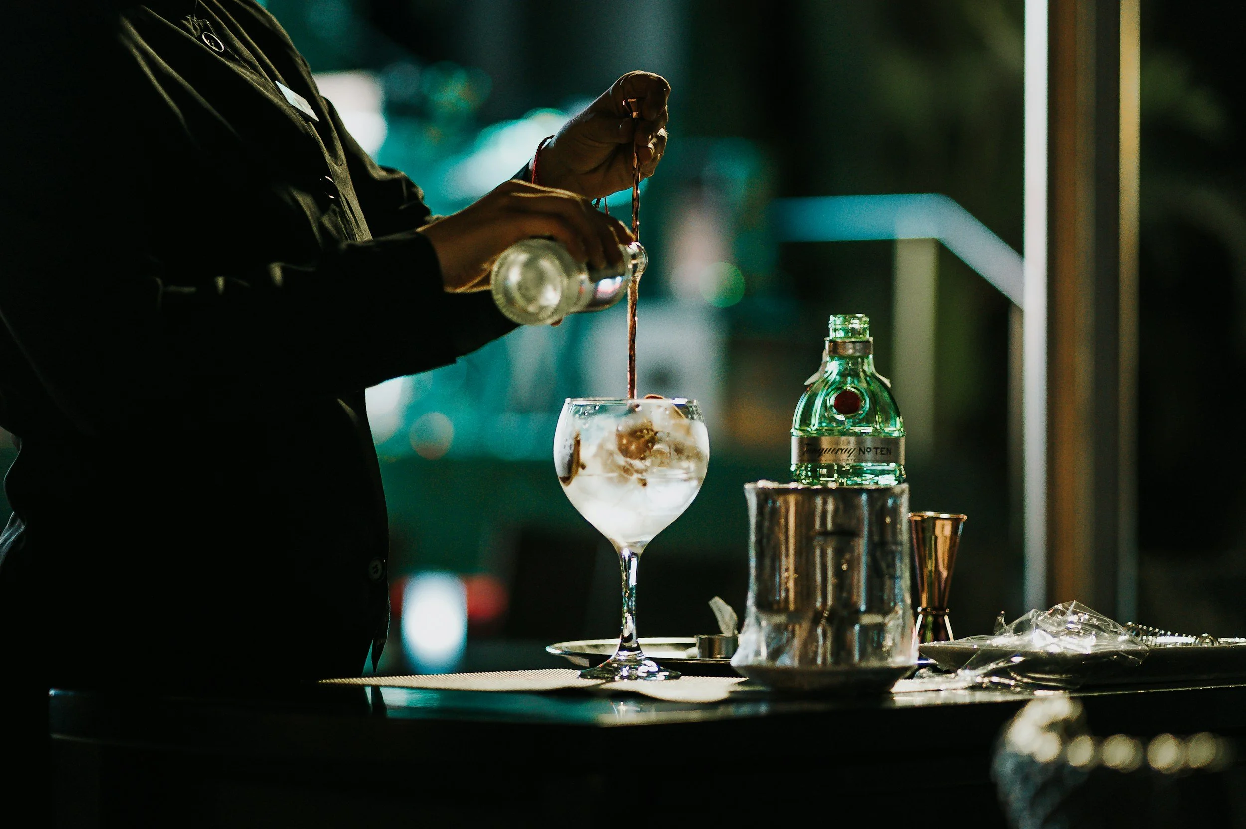 Bartender pouring a drink into a large cocktail glass with ice and soda; bar setup with liquor bottle, ice bucket, and bar tools.