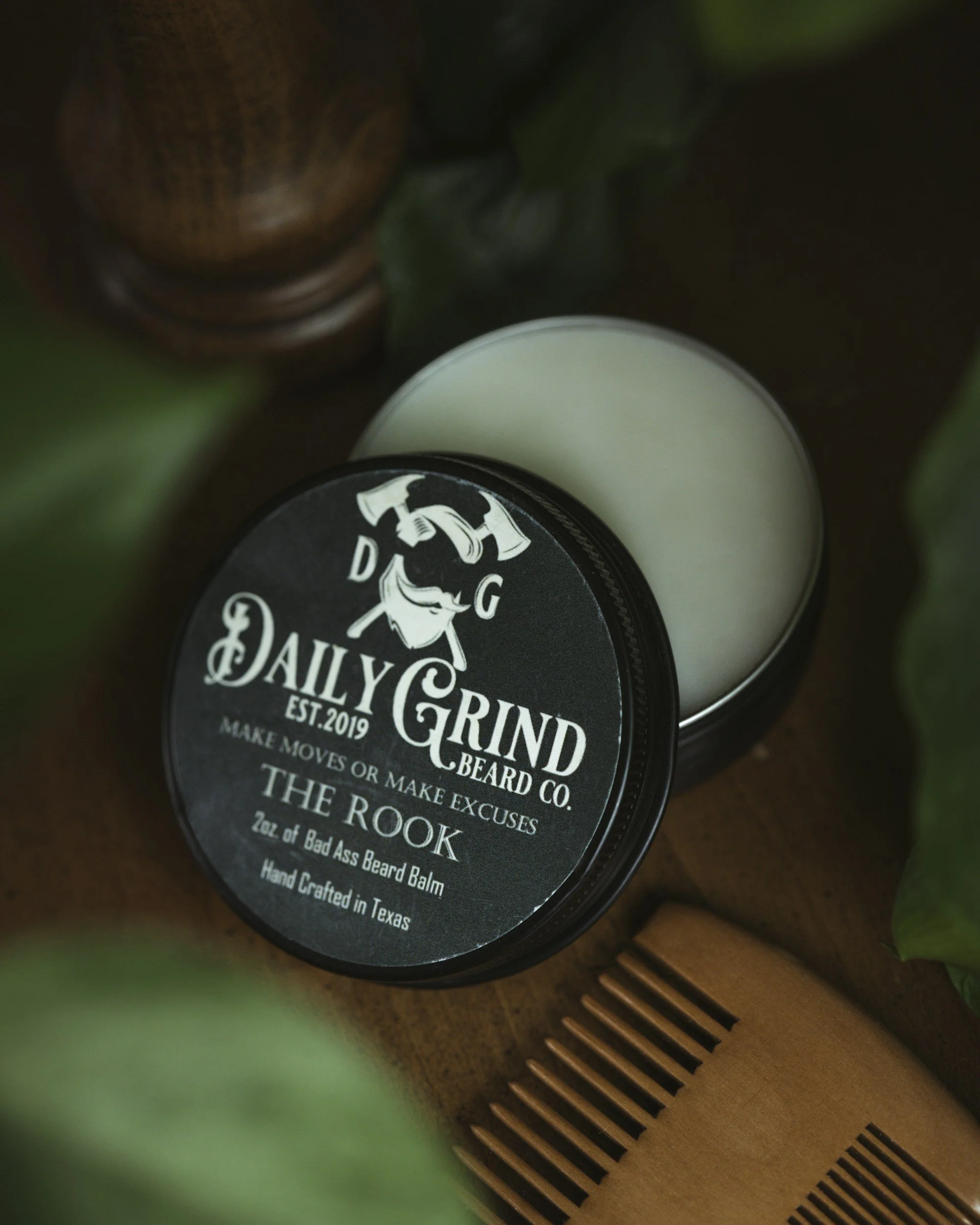 A round tin labeled 'Daily Grind Beard Co.' with a black and white logo of a bearded man and text, placed on a wooden surface with green leaves and a wooden fork nearby.