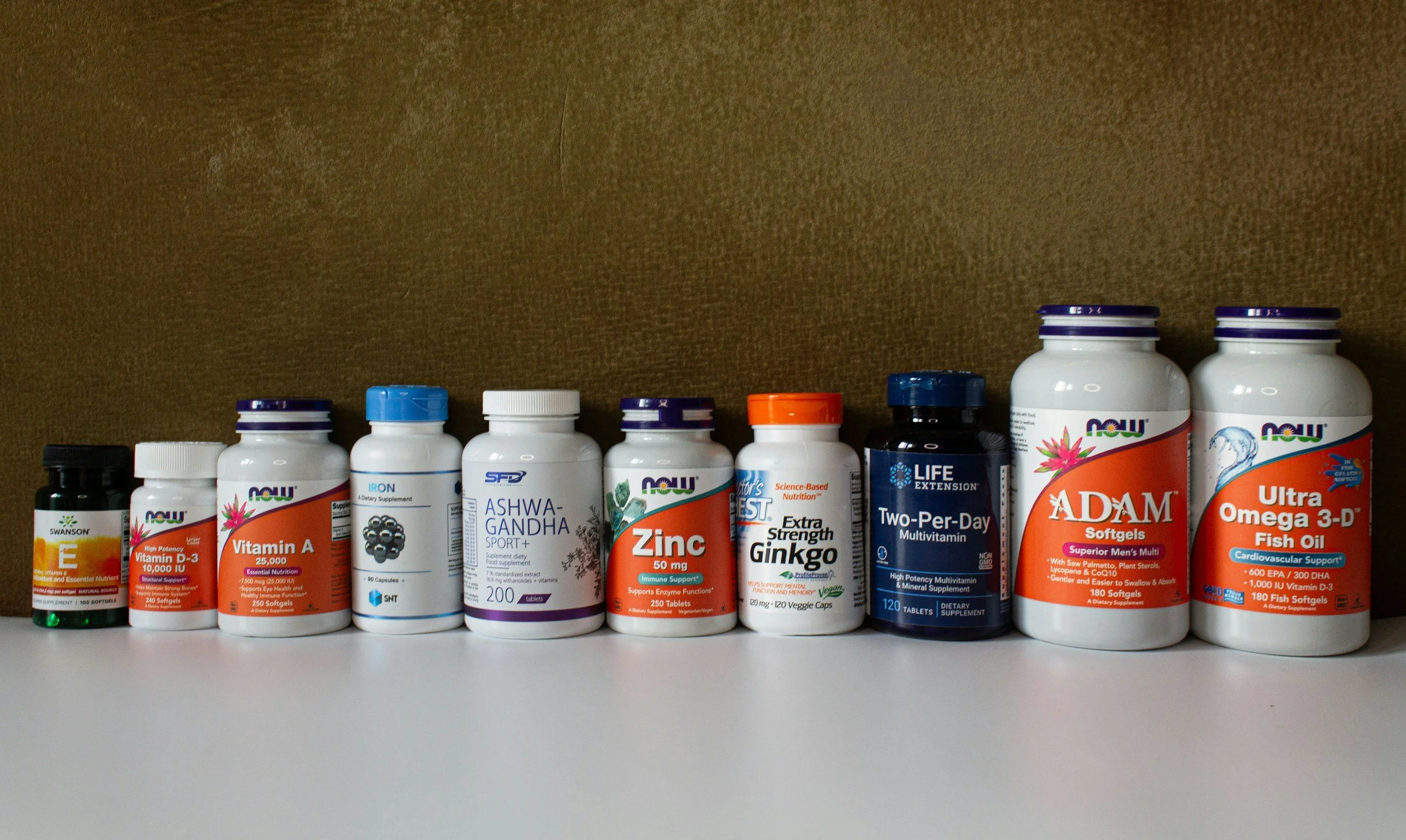 Various dietary supplement bottles, including vitamins and fish oil, arranged on a white surface against a brown wall.