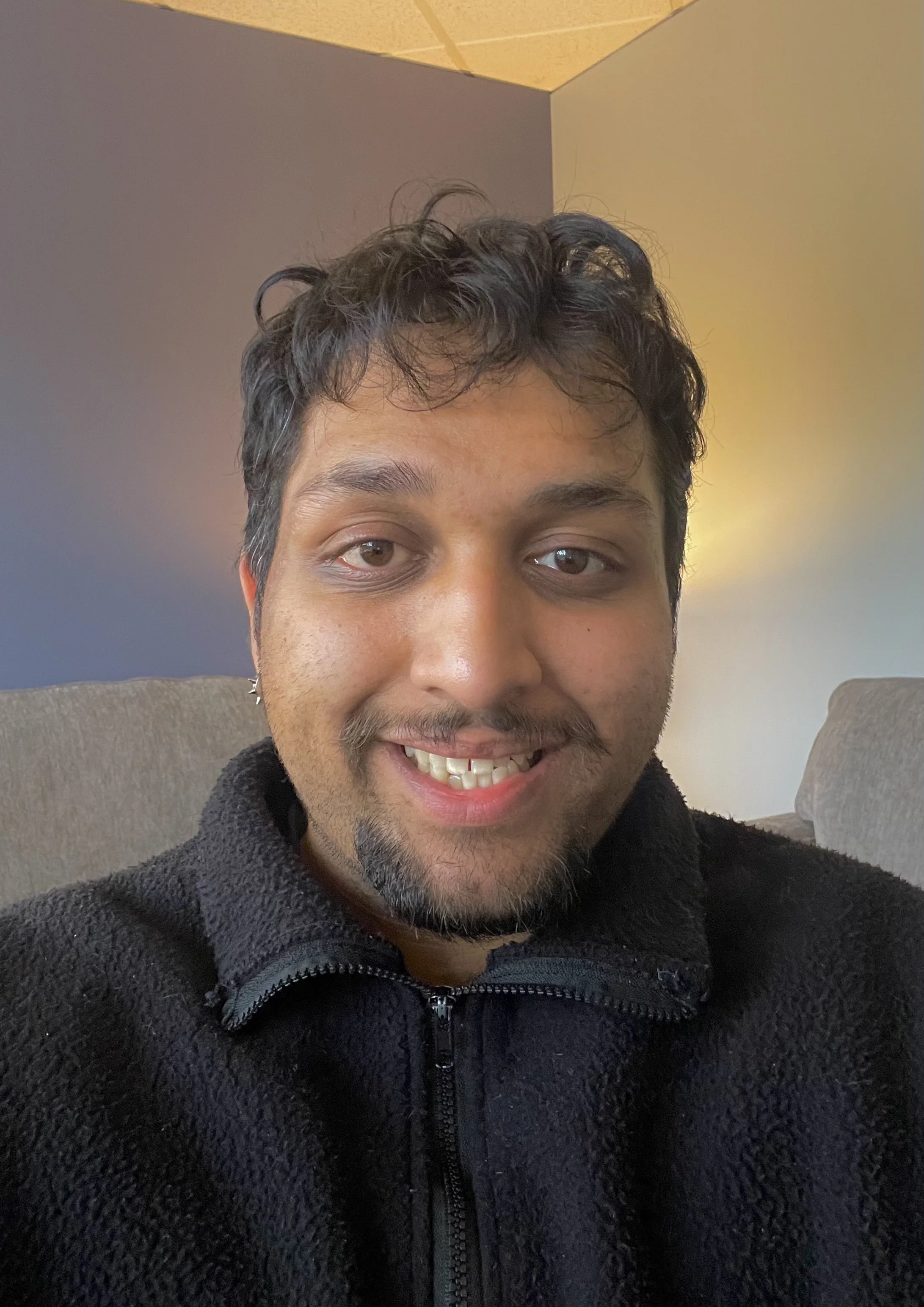 Close-up of a young man with curly dark hair, light skin, and a goatee, smiling at the camera. He is wearing a black fleece jacket, seated on a sofa, with a background of a gray wall with warm ambient lighting.
