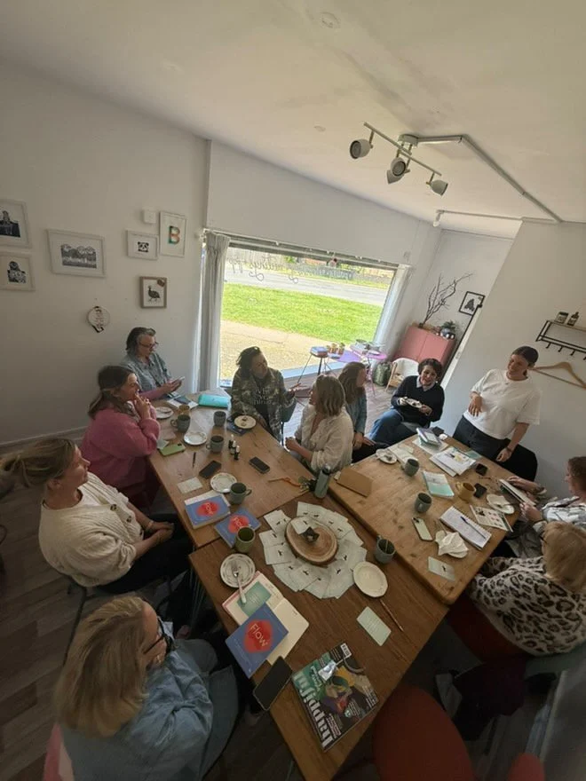 People sitting around a large dining table in a cozy room, engaged in a group discussion or meeting, with various food and beverages on the table, and a large window showing a grassy outdoor area.