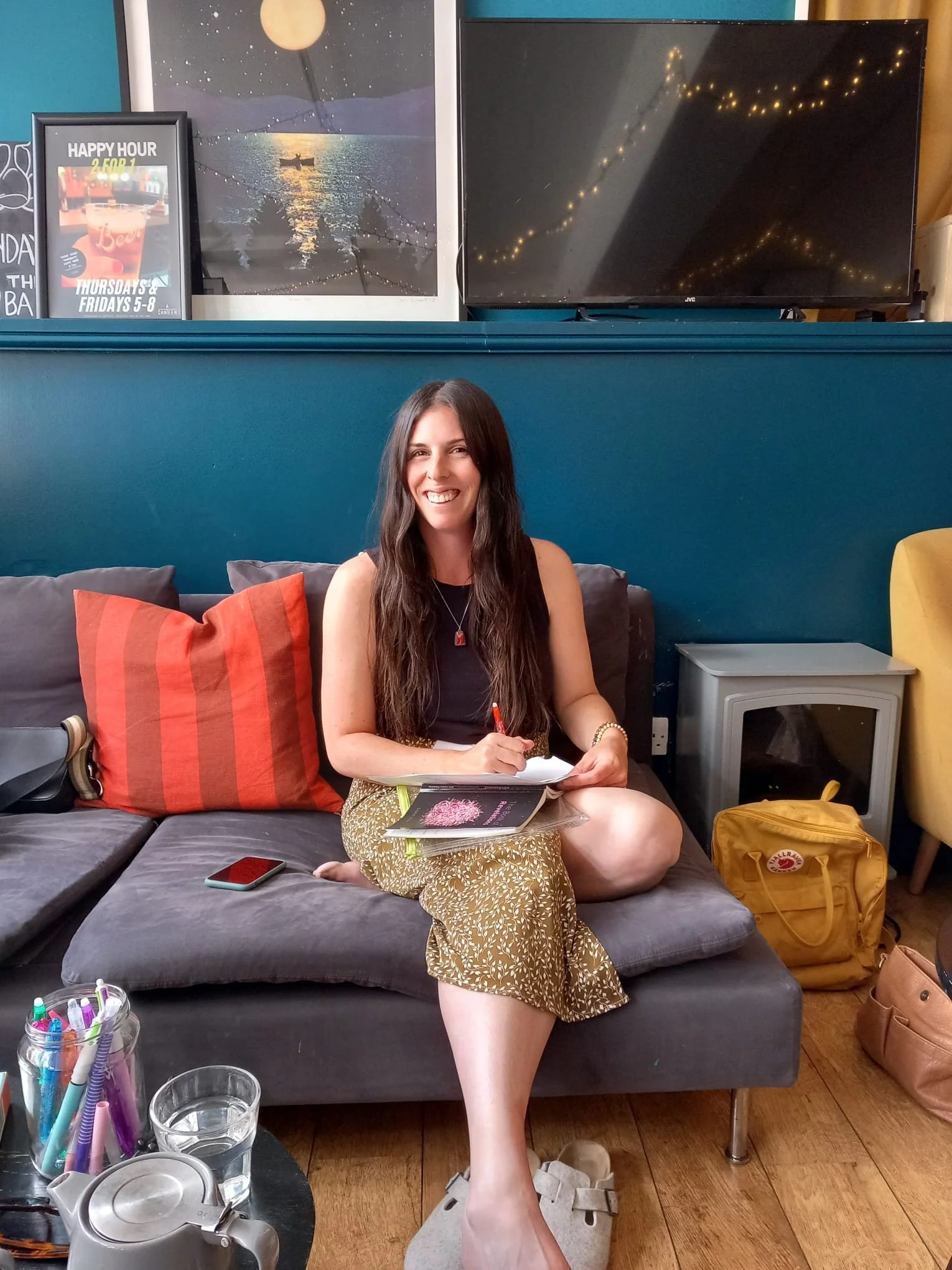 A woman with long dark hair sitting on a grey sofa in a casual indoor setting, smiling and writing on a notebook, with colorful pillows, a remote control, a smartphone, and a glass of water nearby. Behind her are framed pictures, a large TV, and decorative lighting.