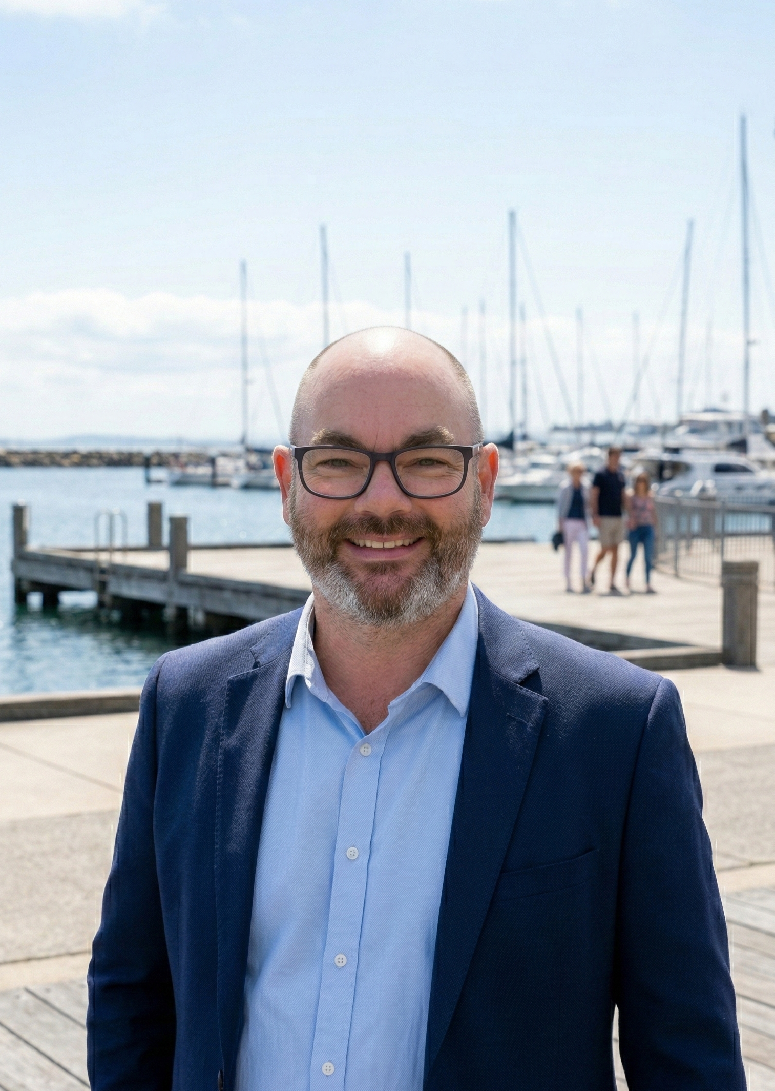 Ben from RoadSafe Driver Training, smiling with glasses and a beard in a navy blazer and light blue shirt standing on a dock near boats in a Geelong marina.