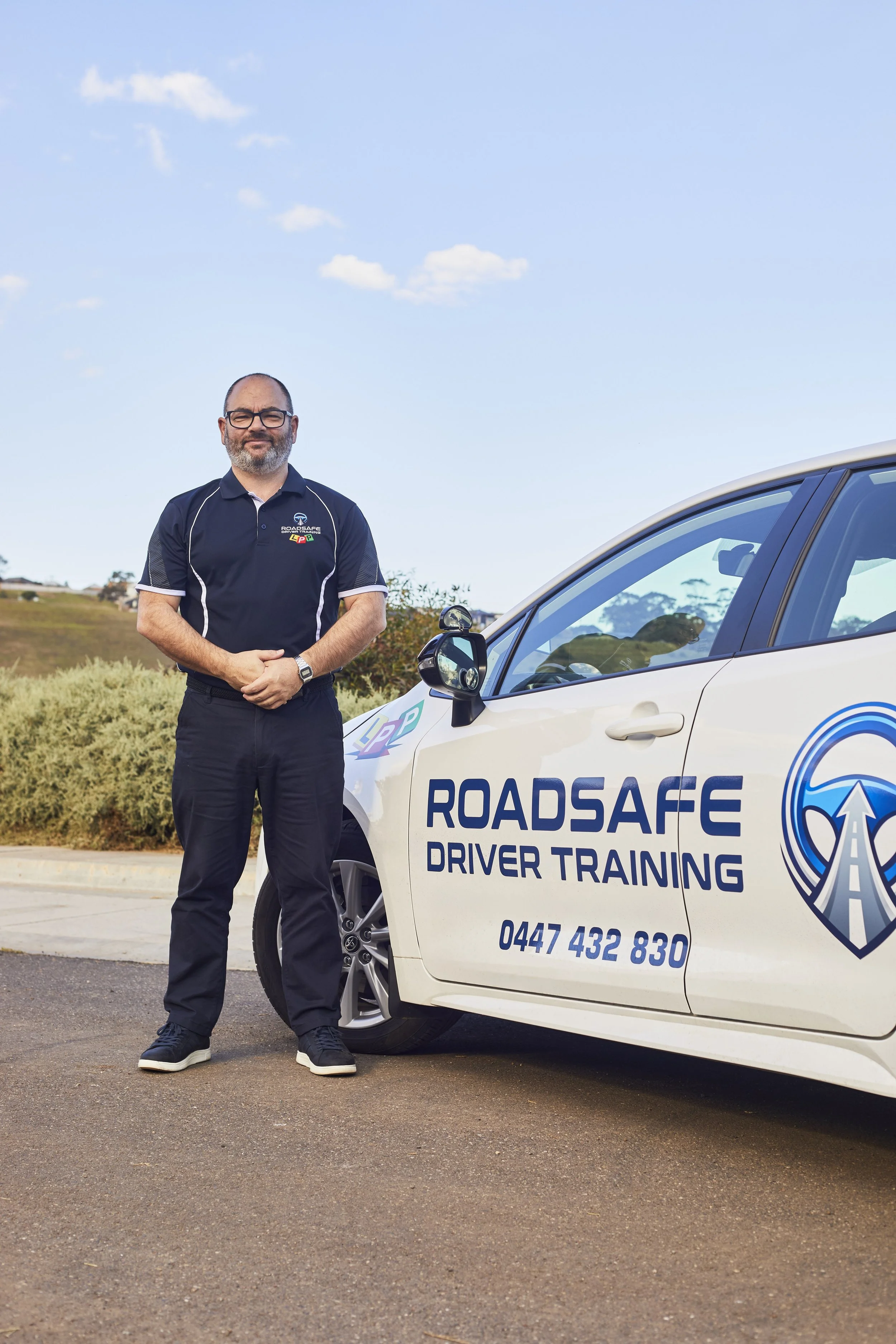 Geelong Driving Instructor 'Roadsafe Driver Training' logo, stands next to a white driving school car with similar logo and contact number on the door, outdoors with a blue sky and open field in background.