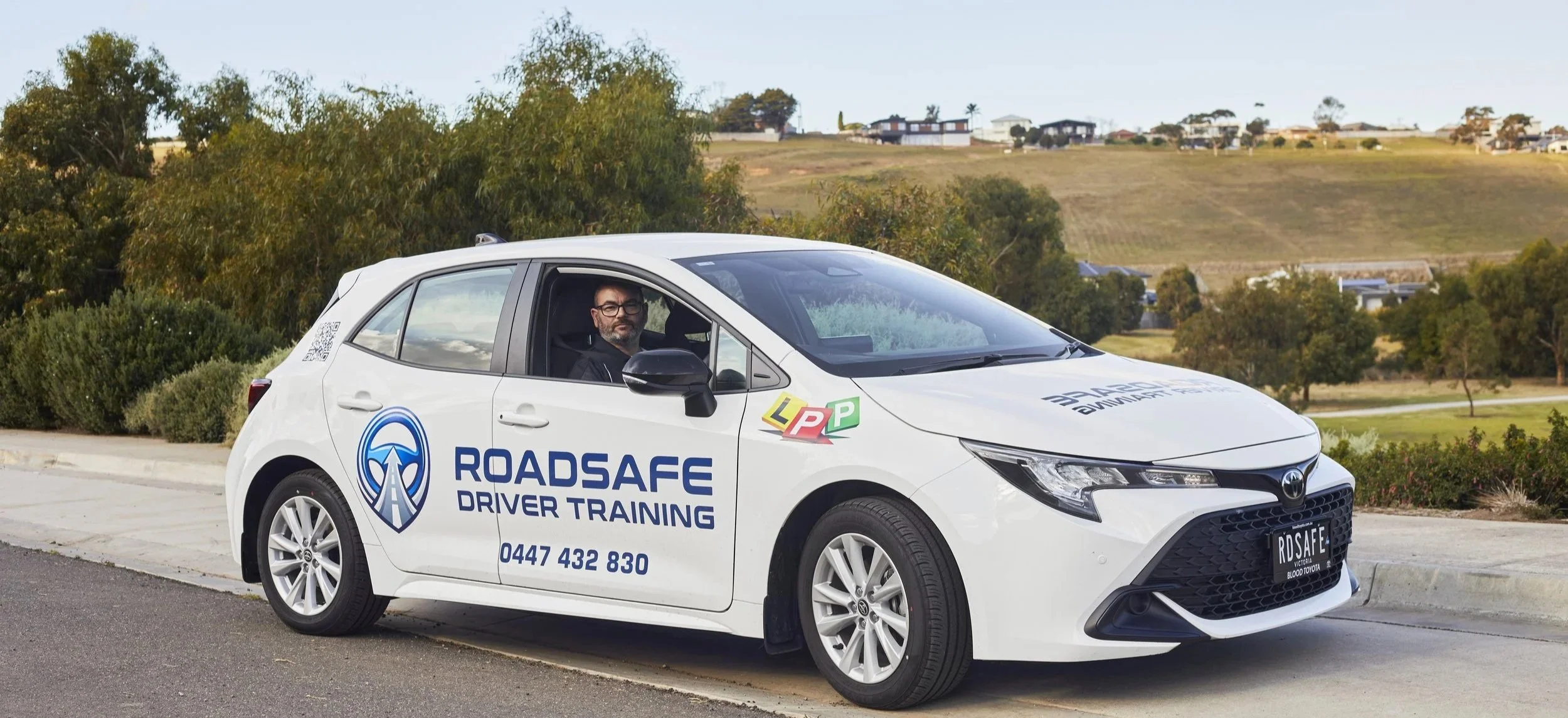 A man sitting in the driver's seat of a white car with 'RoadSafe Driver Training' branding, parked on a road with green trees and hills in the background.