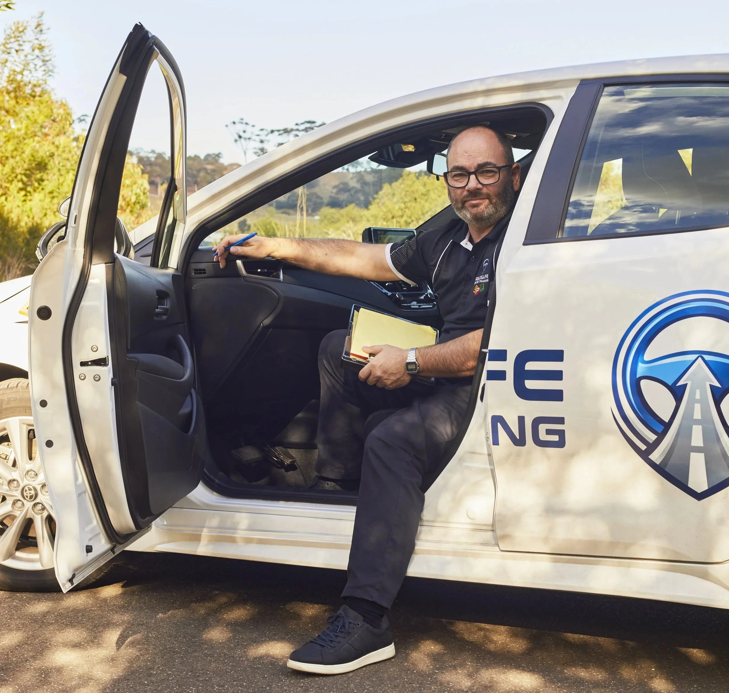 A man with glasses and a beard sitting in the driver's seat of a white vehicle, possibly a police car, holding a yellow notepad and pen, with the driver's side door open, outdoors during daytime with trees and a partly cloudy sky in the background.