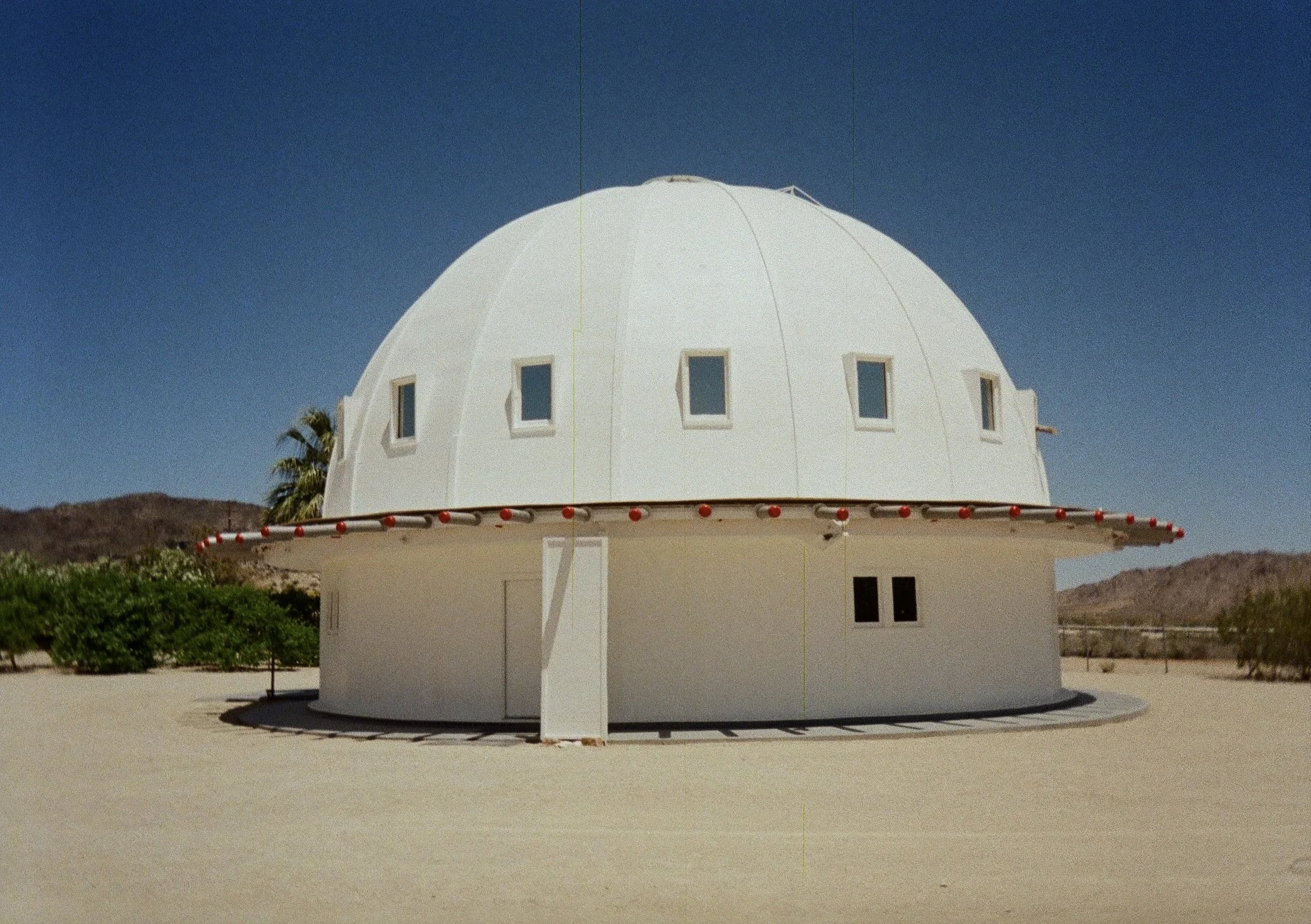 White dome-shaped building with small rectangular windows, red lights around the roof, set in a desert landscape under a clear blue sky.