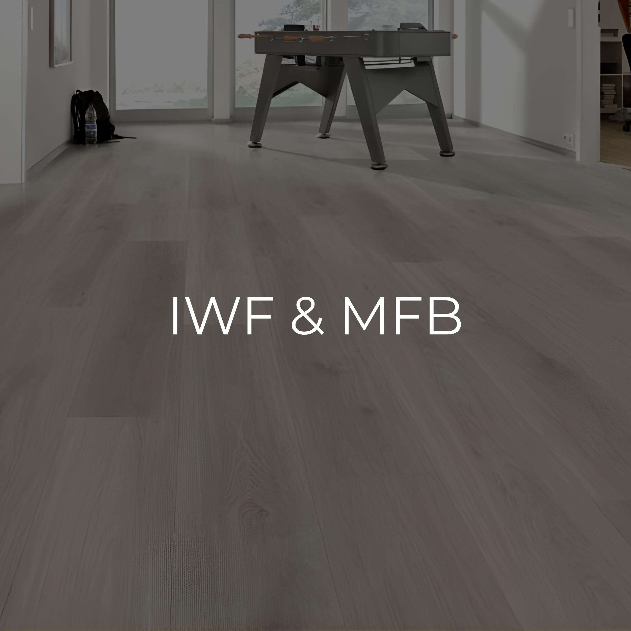 Interior view of a room with light-colored wooden flooring, a foosball table, and a large window with a view outside, with text overlay reading "IWF & MFB."