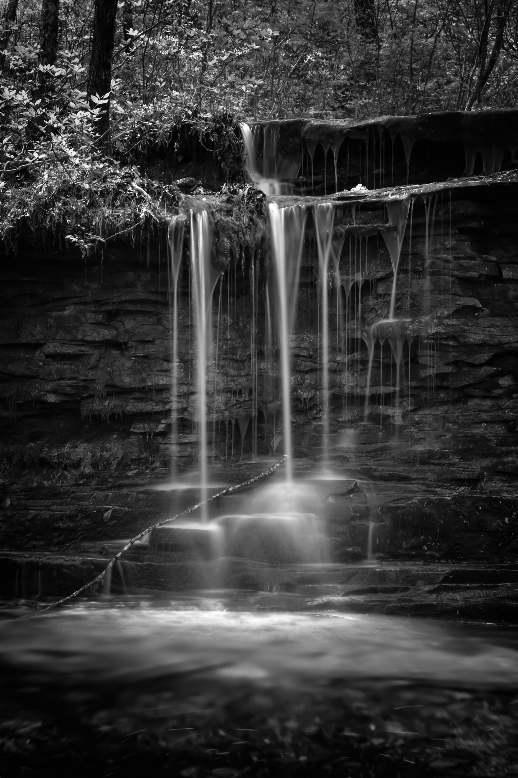 Black and white long exposure of gently illuminated multi-tiered waterfall flowing over mossy rock ledge into calm forest pool surrounded, DeSoto State Park, Alabama.