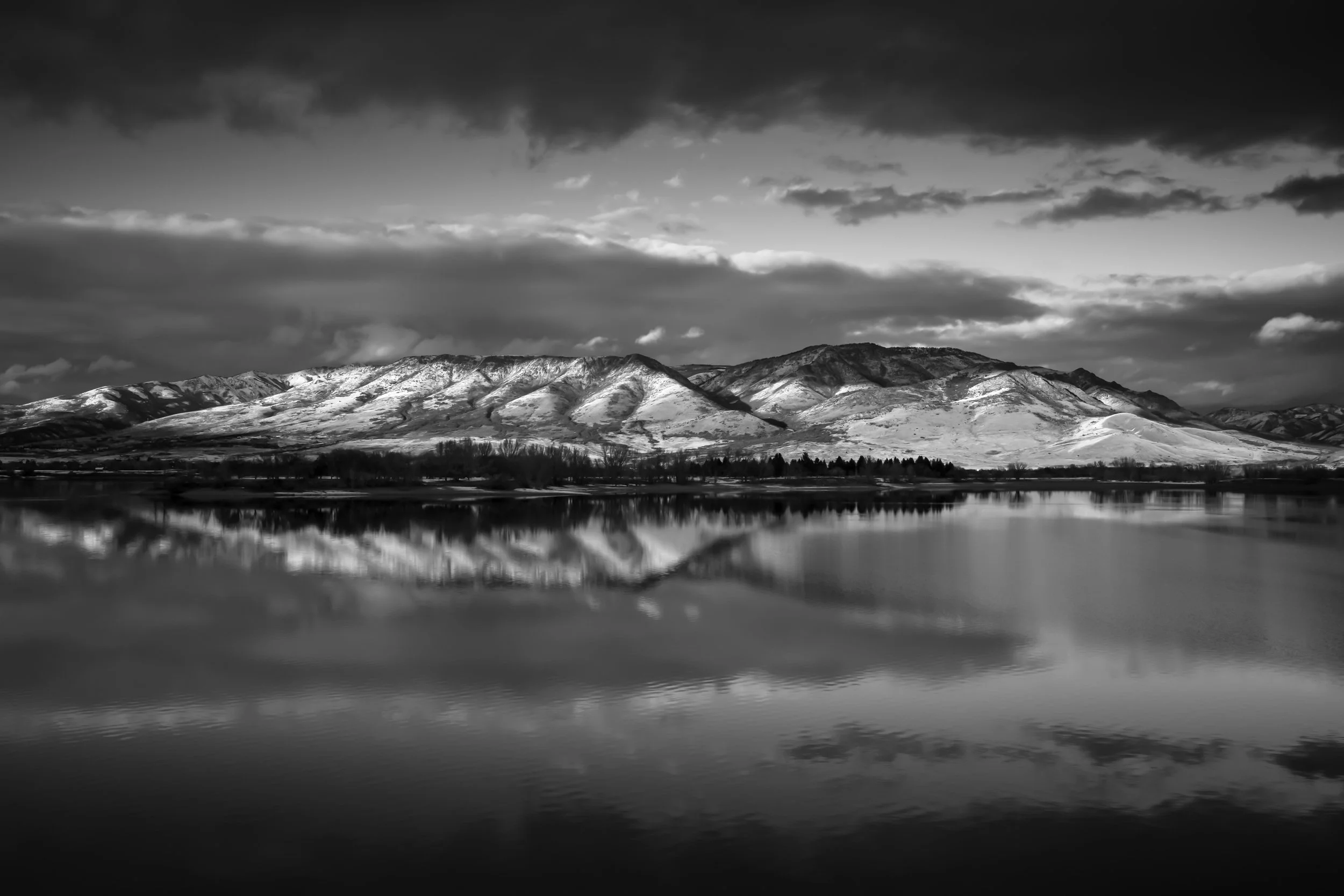 Dramatic black and white landscape of mountain range reflected in still water under moody cloudy sky, Pineview Reservoir, Ogden Valley, Utah.