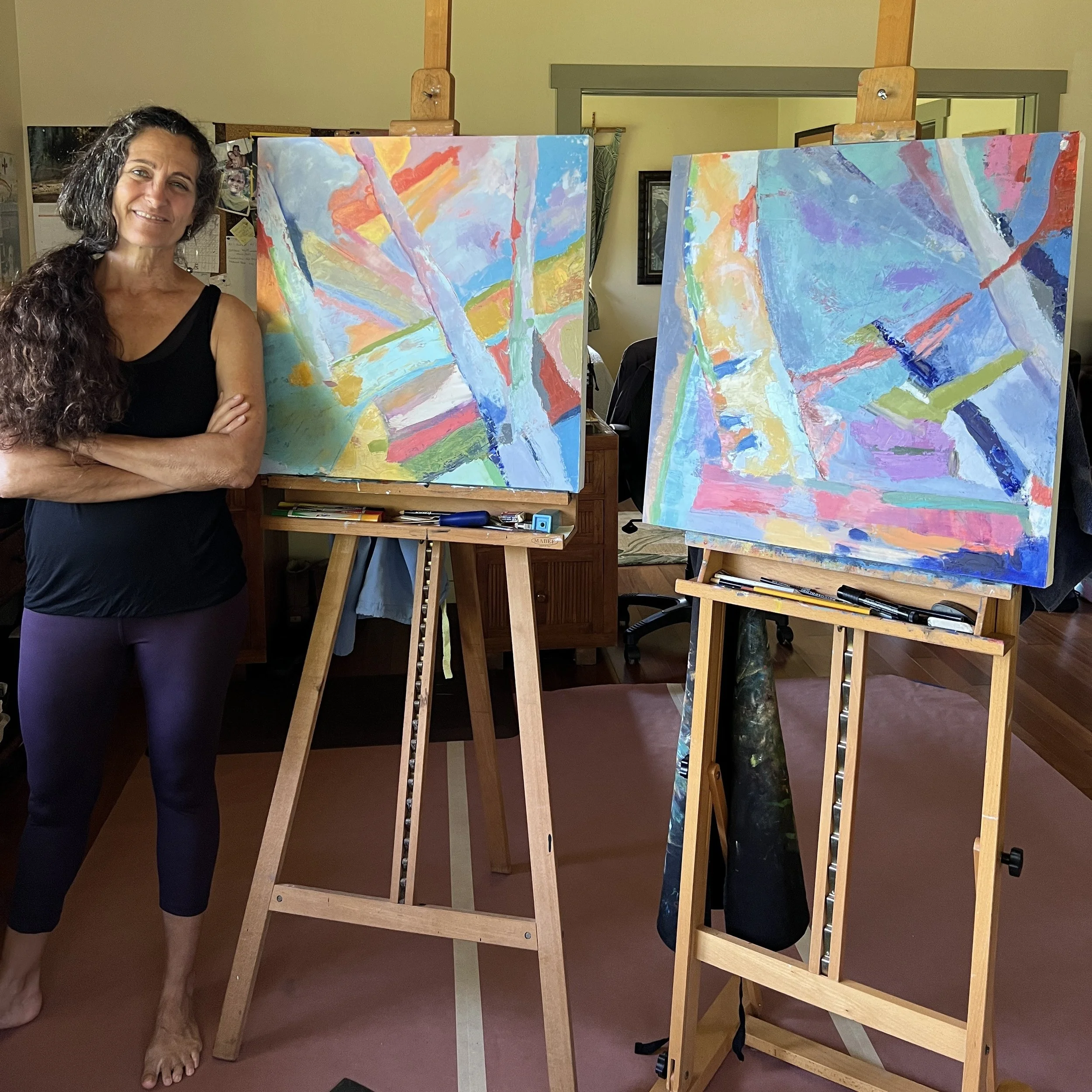 A woman with long dark curly hair and a black sleeveless top standing with arms crossed next to two colorful abstract paintings on easels in an indoor studio.