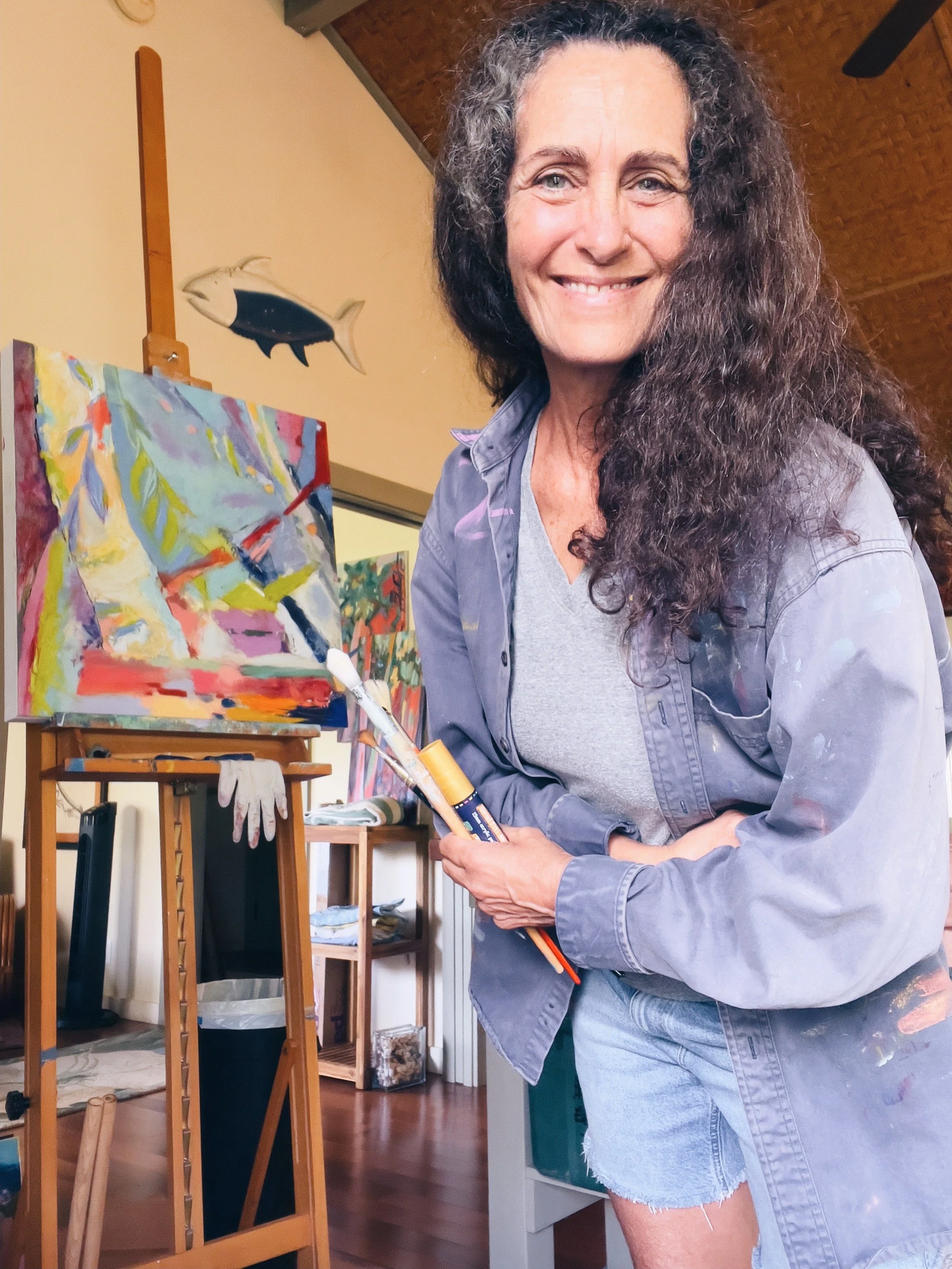 A woman with long curly hair smiling while holding paintbrushes and standing next to an easel with an abstract painting, in a room with a fish wall decoration
