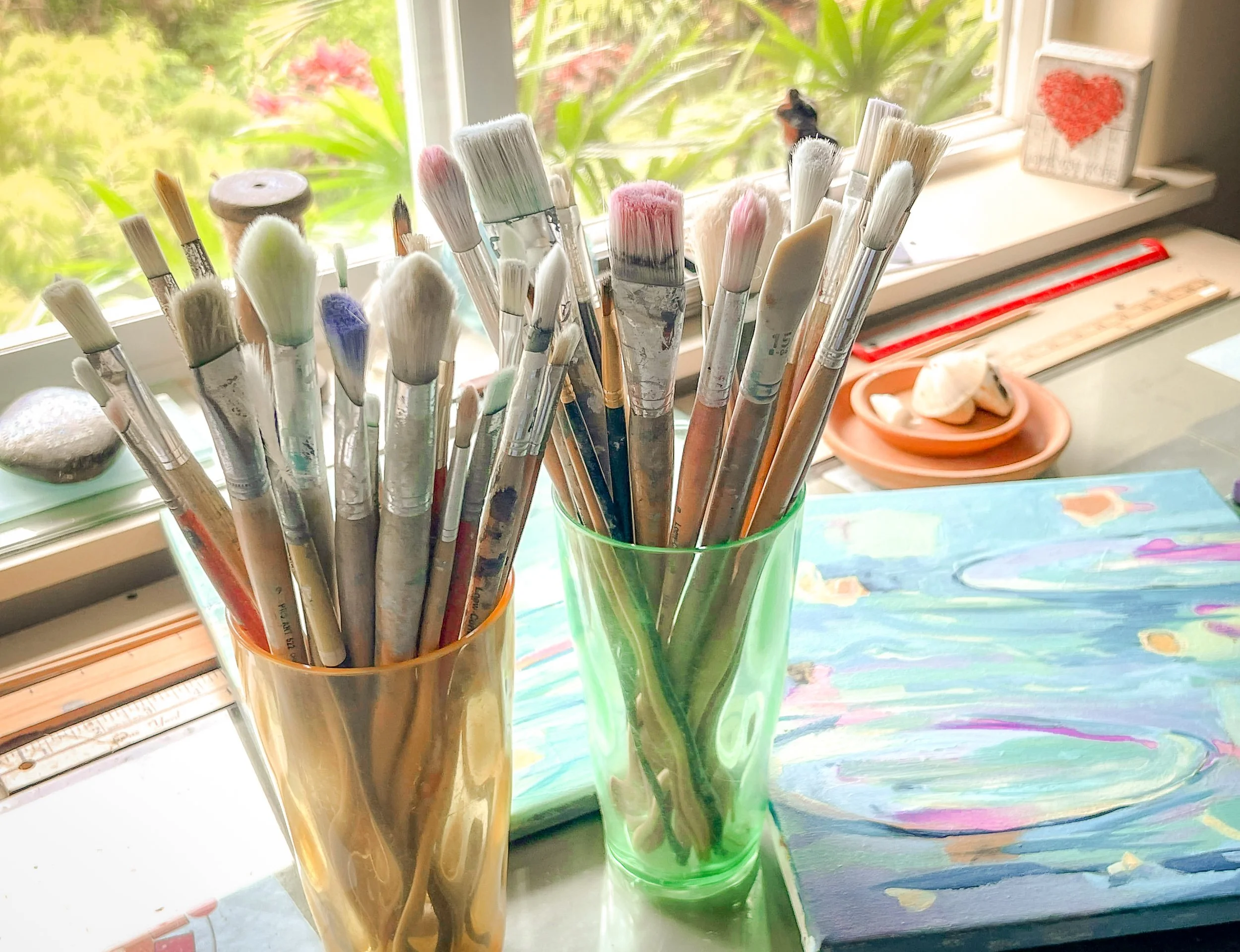 Two cups filled with various paintbrushes sit on an artist's workspace near a window, with a colorful painting and small art supplies in the background.
