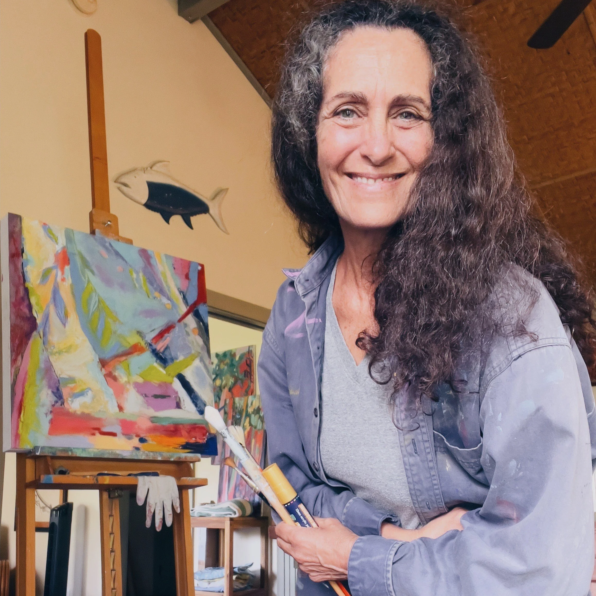 A woman with long curly gray hair smiling and holding paintbrushes stands next to an abstract painting on an easel in a cozy indoor space.
