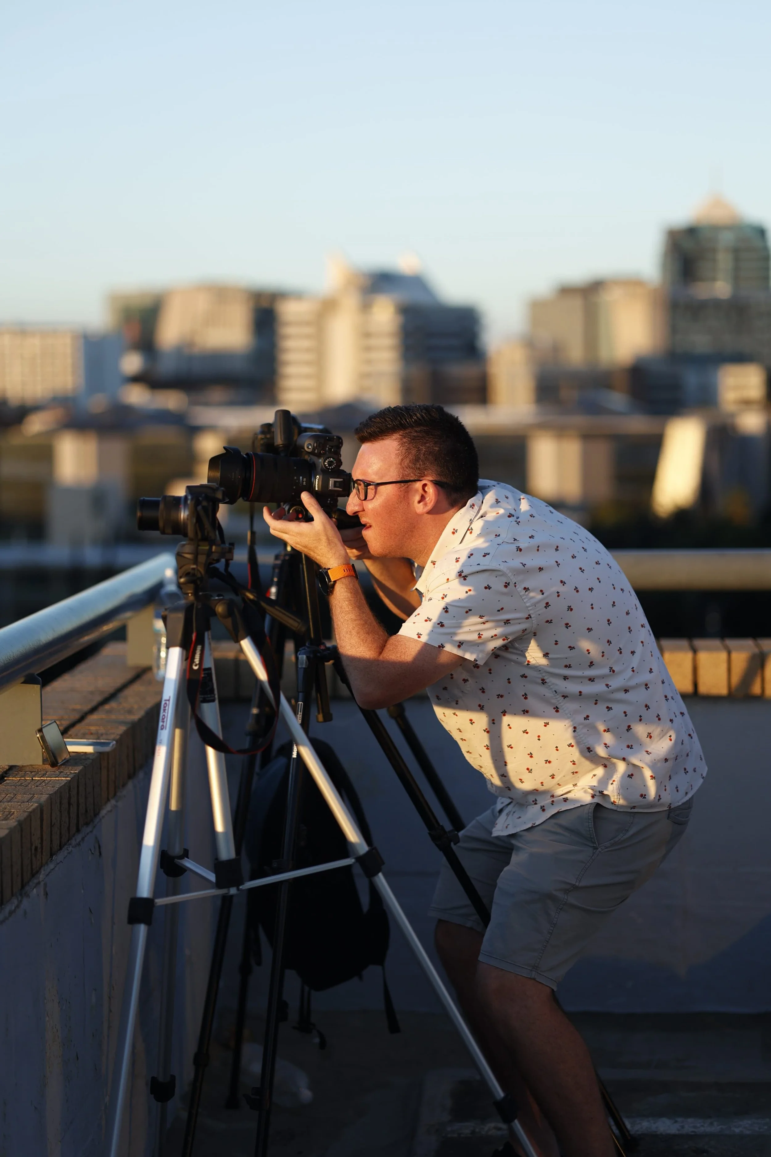 A man wearing glasses and a white shirt with small floral prints is taking photos with a camera mounted on a tripod on a rooftop during sunset, with an urban cityscape in the background.