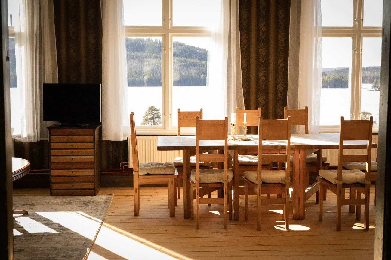 Vy av ett matsalsrum med ett stort bord, åtta trästolar med sitskuddar, fönster som visar en snötäckta landskap utomhus, en TV på en träskänk, ljus insläpp, och gardiner.
