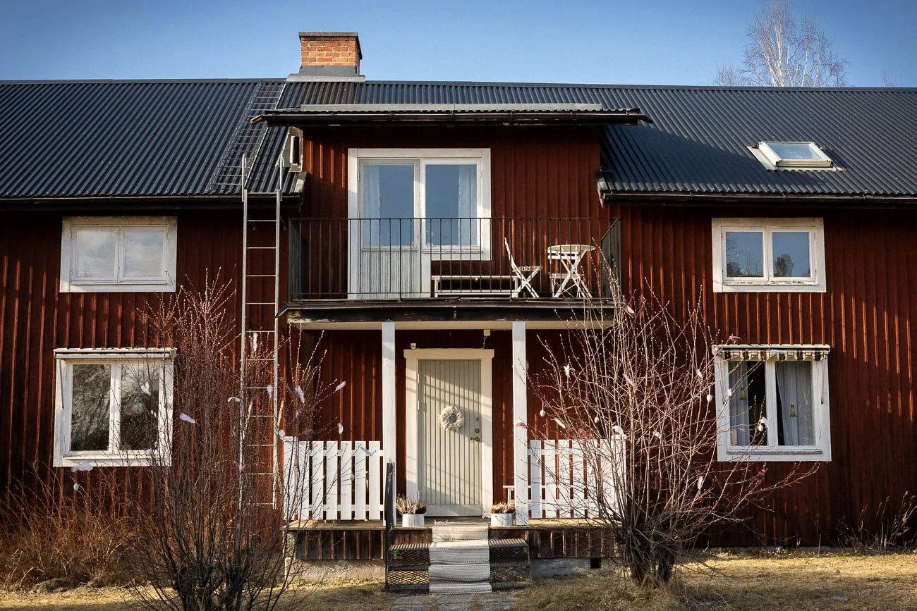 En röd trähus med vita fönster och svart tak, utomhus med buskar och en liten stig framför entrén.