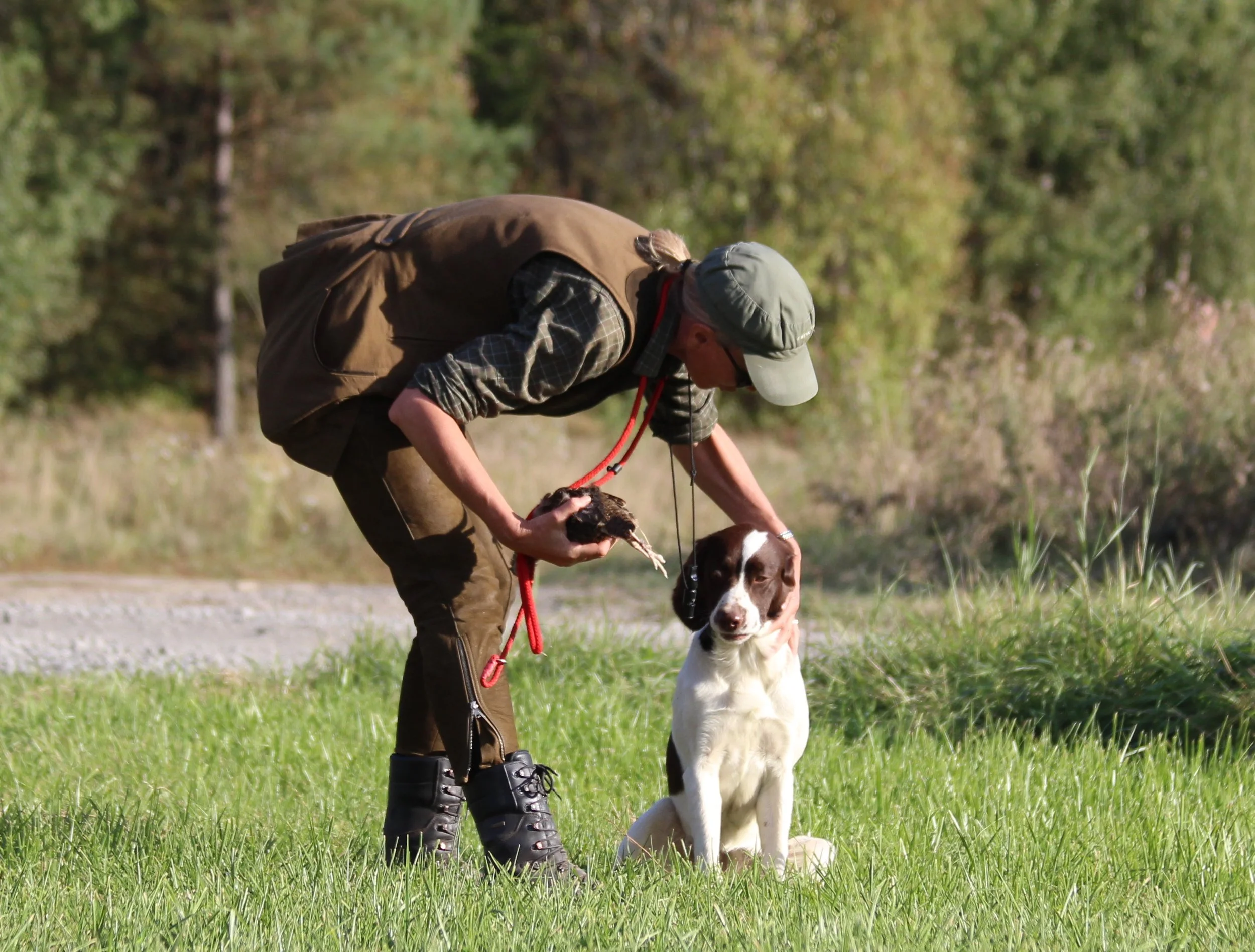Person i utomhusmiljö med hund, håller i en fågel, grönt gräs och träd i bakgrunden.