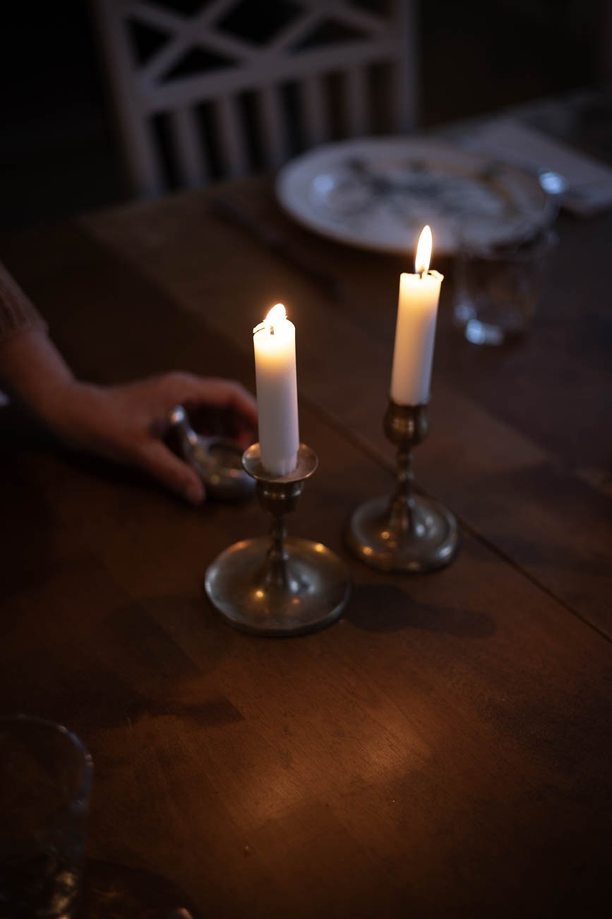 Två vita stearinljus i bronsfärgade ljusstakar står på ett mörkt trähambord i ett dukat matsalsrum.