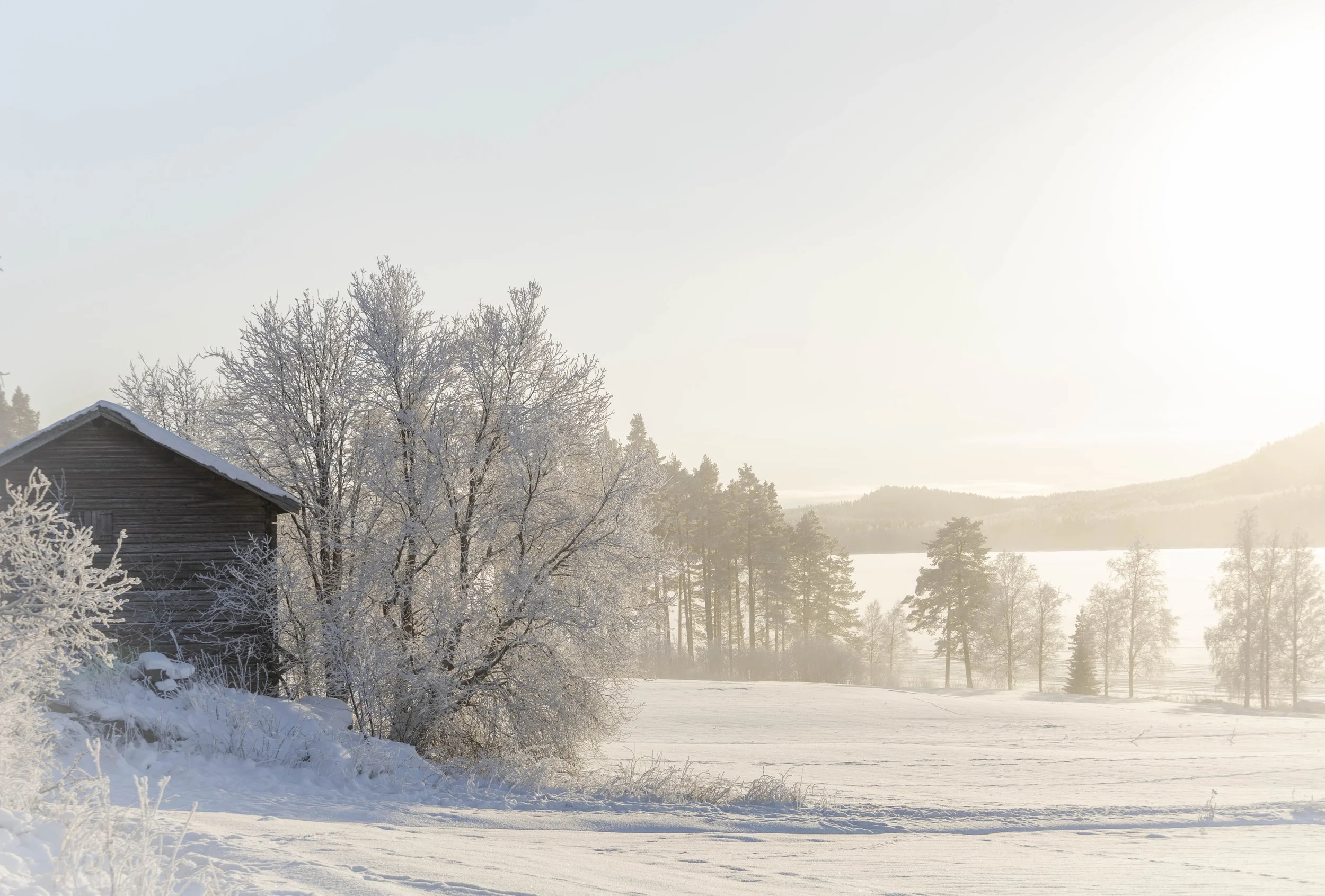 Snötäckta landskap med ett litet hus, snöbeklädda träd och en sjö i bakgrunden under solnedgången.