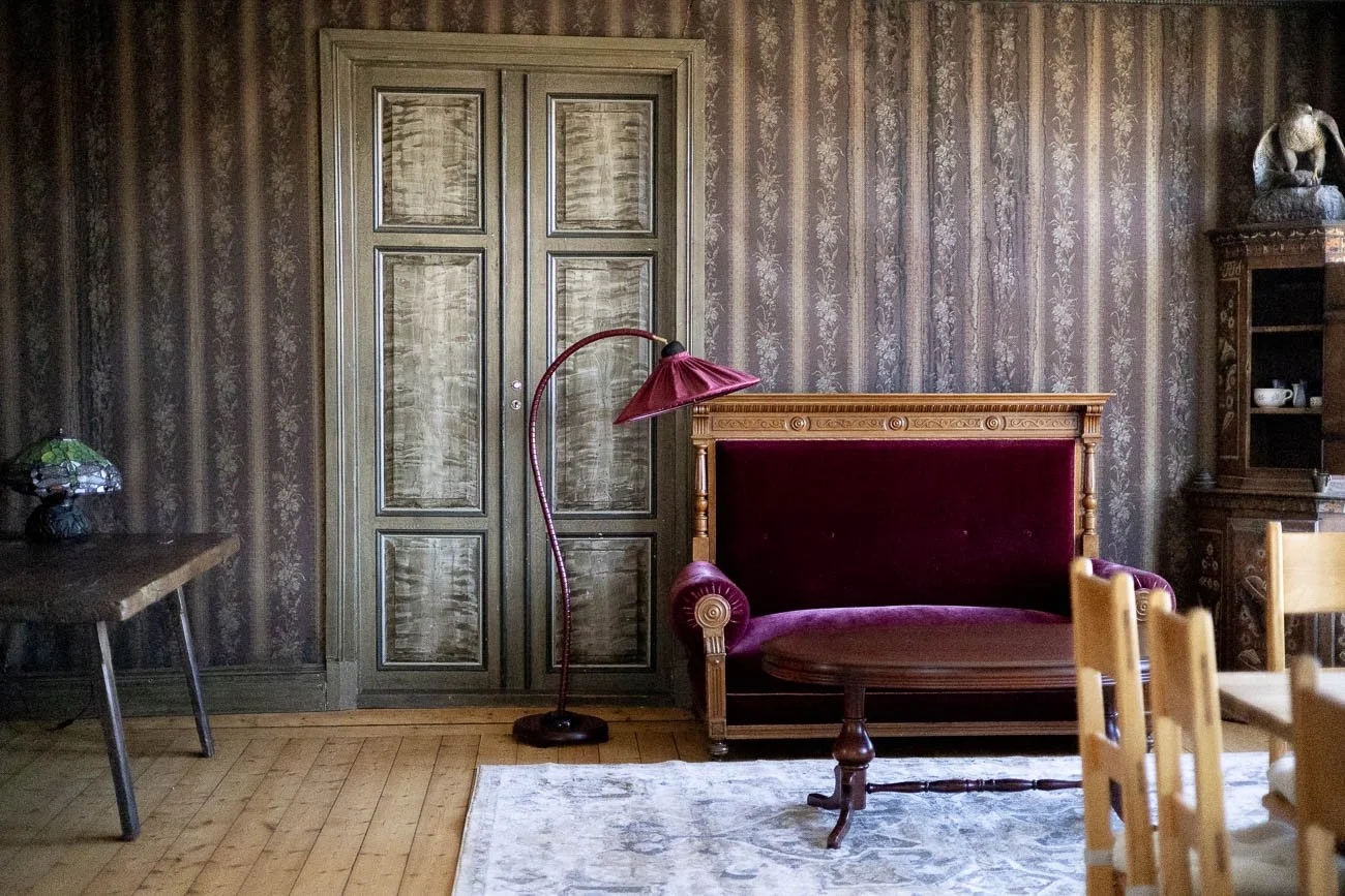 Väggar med vintage tapet, ett skåp, en madrass på en träram, en golvlampa, ett bord och en matta i ett rumm.