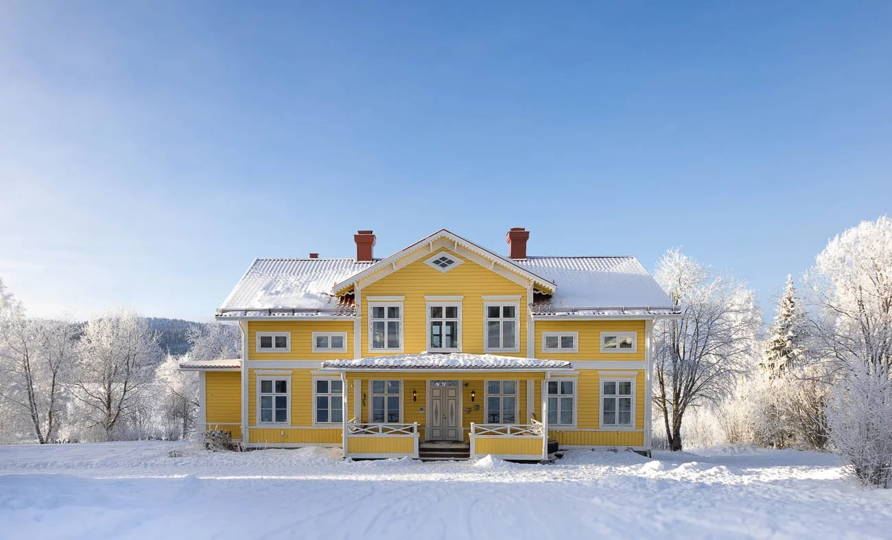 En gul stor hus med vit dörr och vinteromgivningar, snö på marken och frost på träden, under en klar blå himmel.