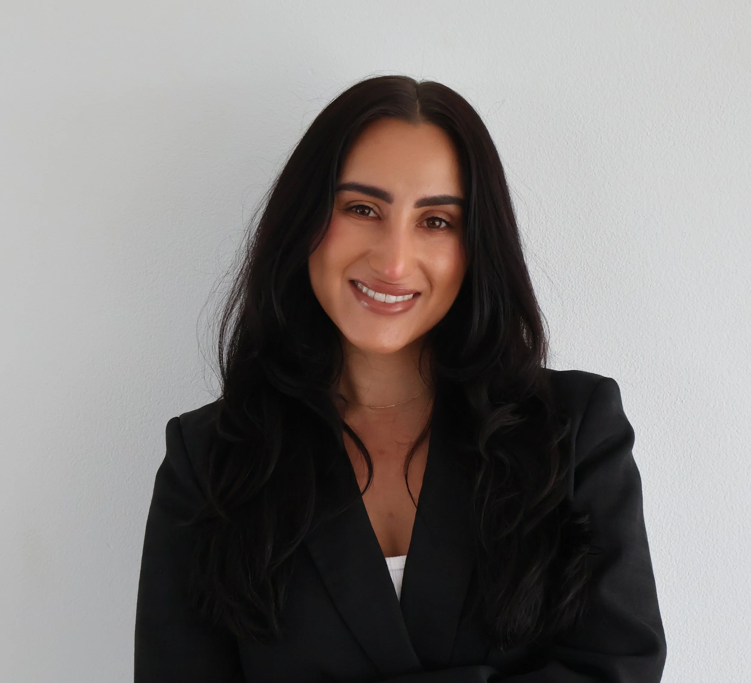 A woman with long dark hair smiling and wearing a black blazer against a plain white wall.