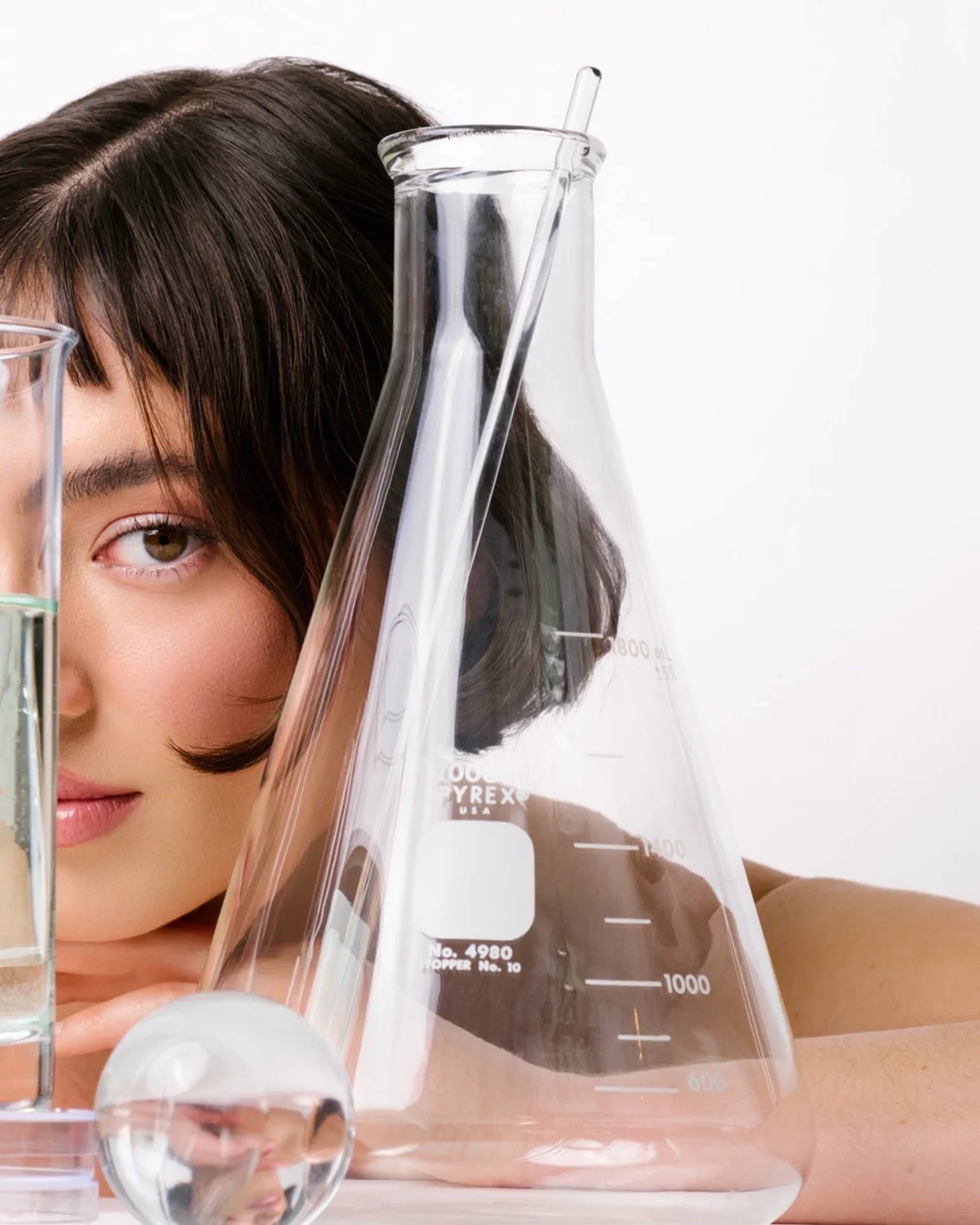 A woman with dark hair and brown eyes surrounded by laboratory glassware, including beakers and an Erlenmeyer flask, with a light background.