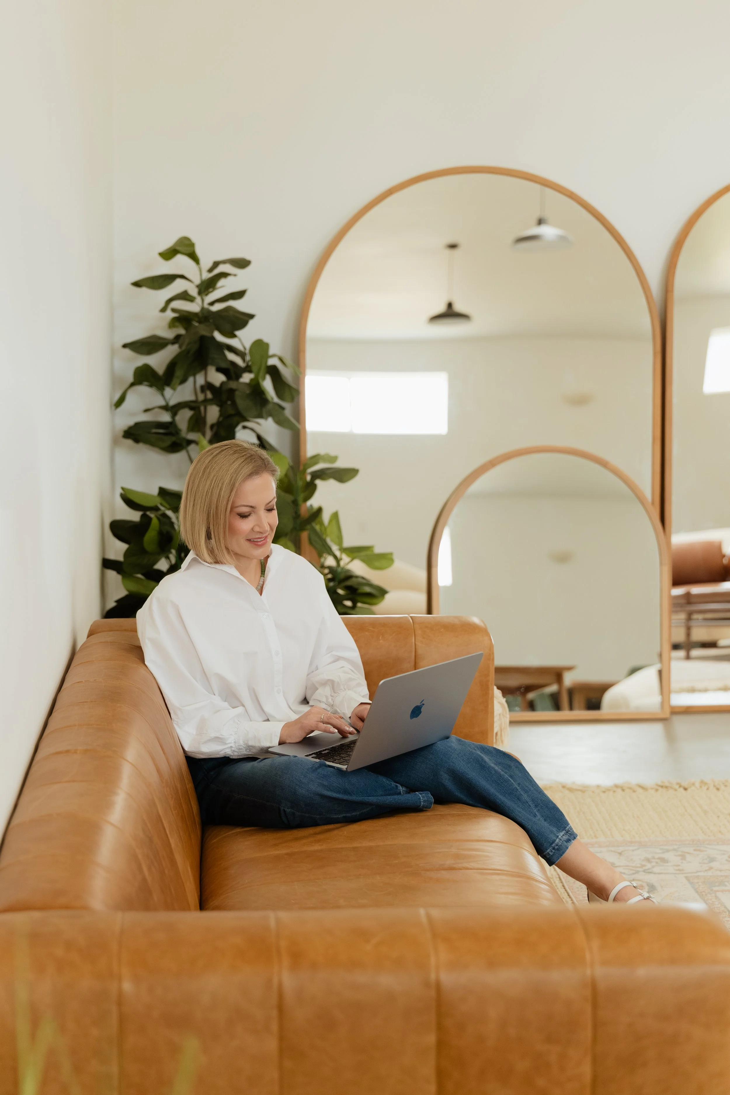 A woman with blonde hair sitting on a brown leather sofa, working on a silver Macbook.