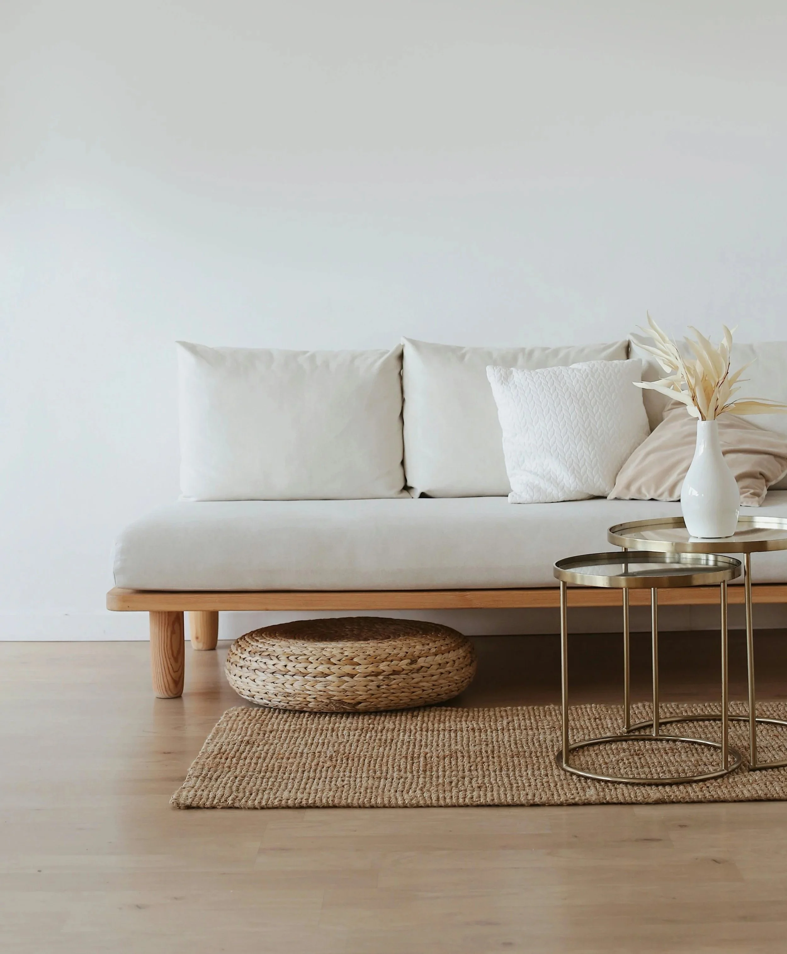 Minimalist living room with a white sofa, neutral cushions, a woven pouf, a jute rug, and round metal side tables with a white vase holding dried leaves.