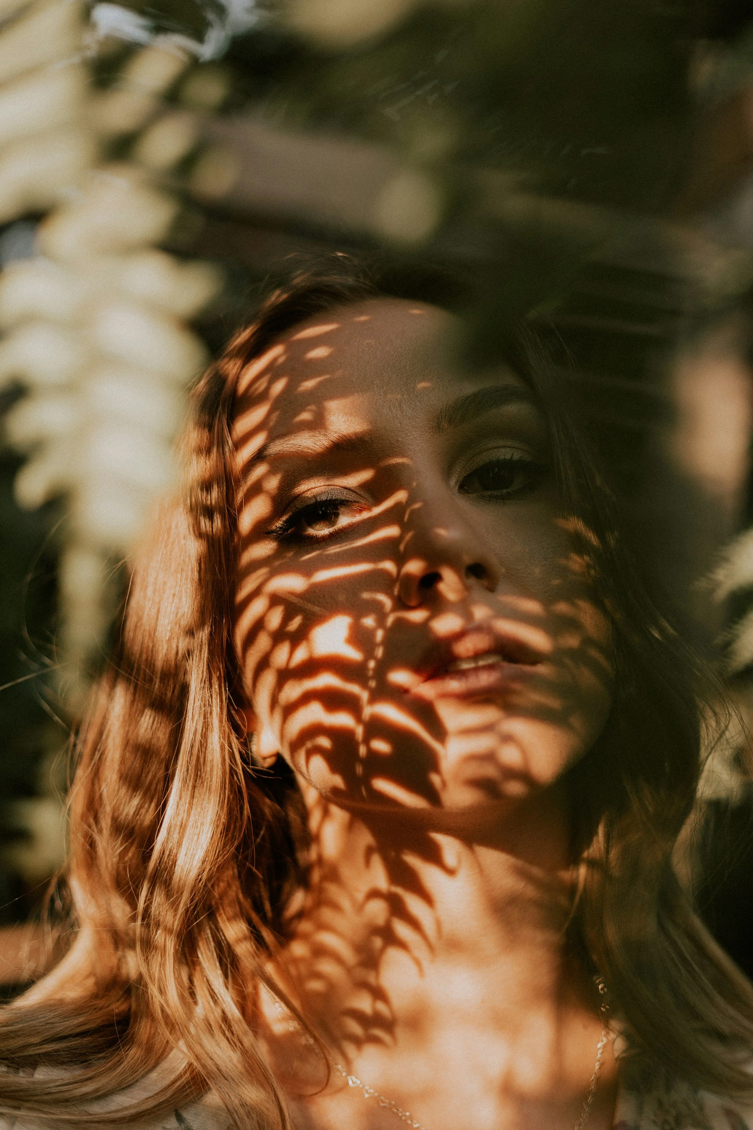 A woman with light brown hair and makeup, partially obscured by shadows cast from leaves, creating a pattern on her face.