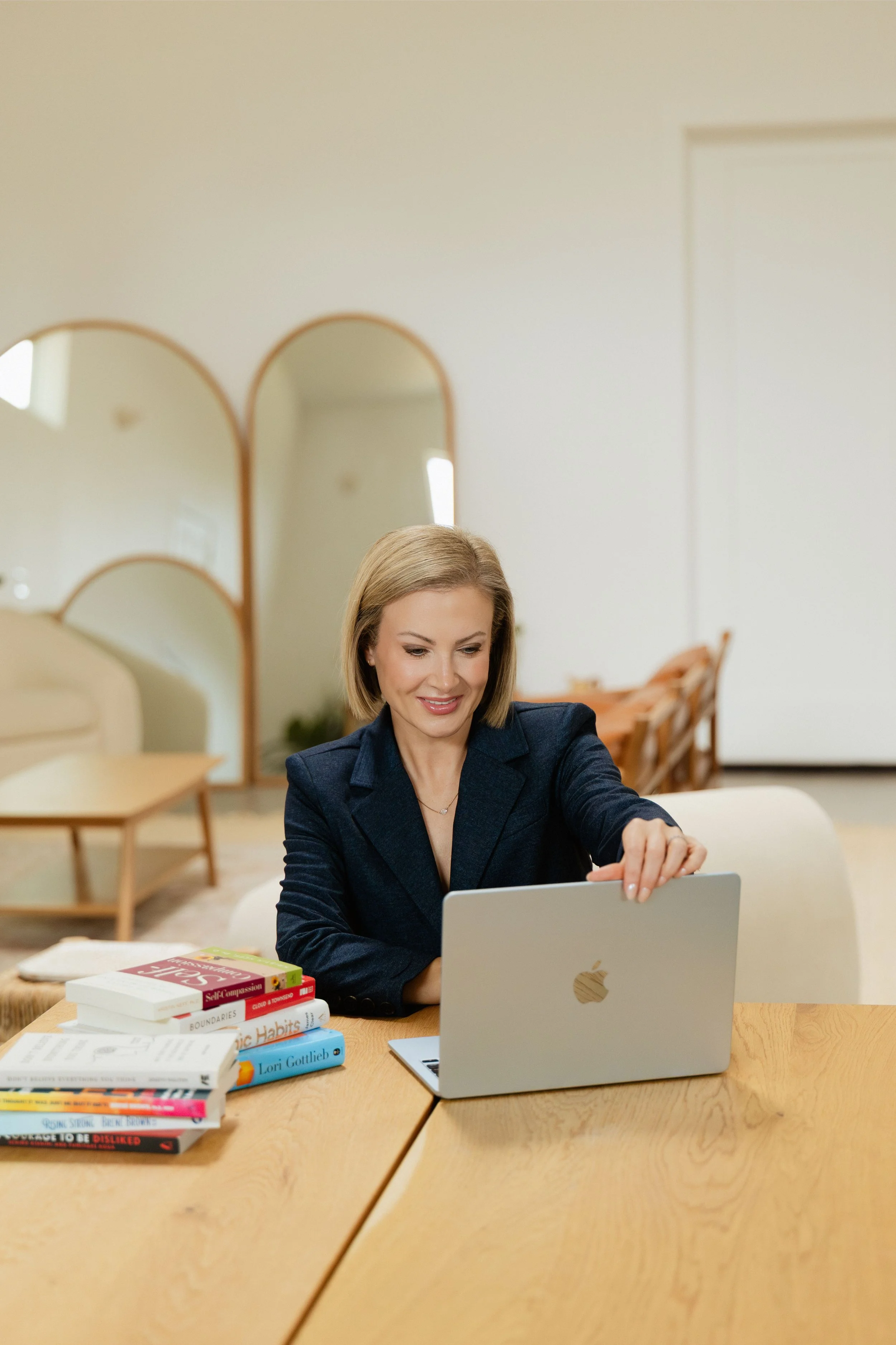 A woman sitting at a wooden table using a silver laptop, with a stack of books nearby, in a bright, modern room with a beige sofa and mirrors in the background.