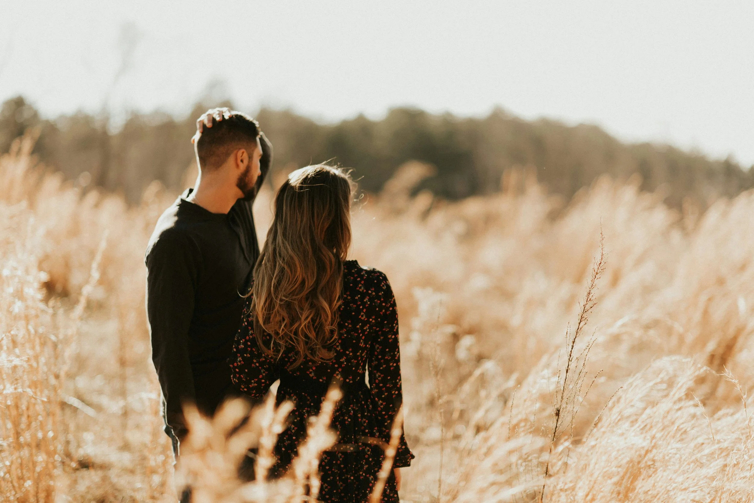 A man and woman standing together in a field of tall, golden grass, facing away from the camera.