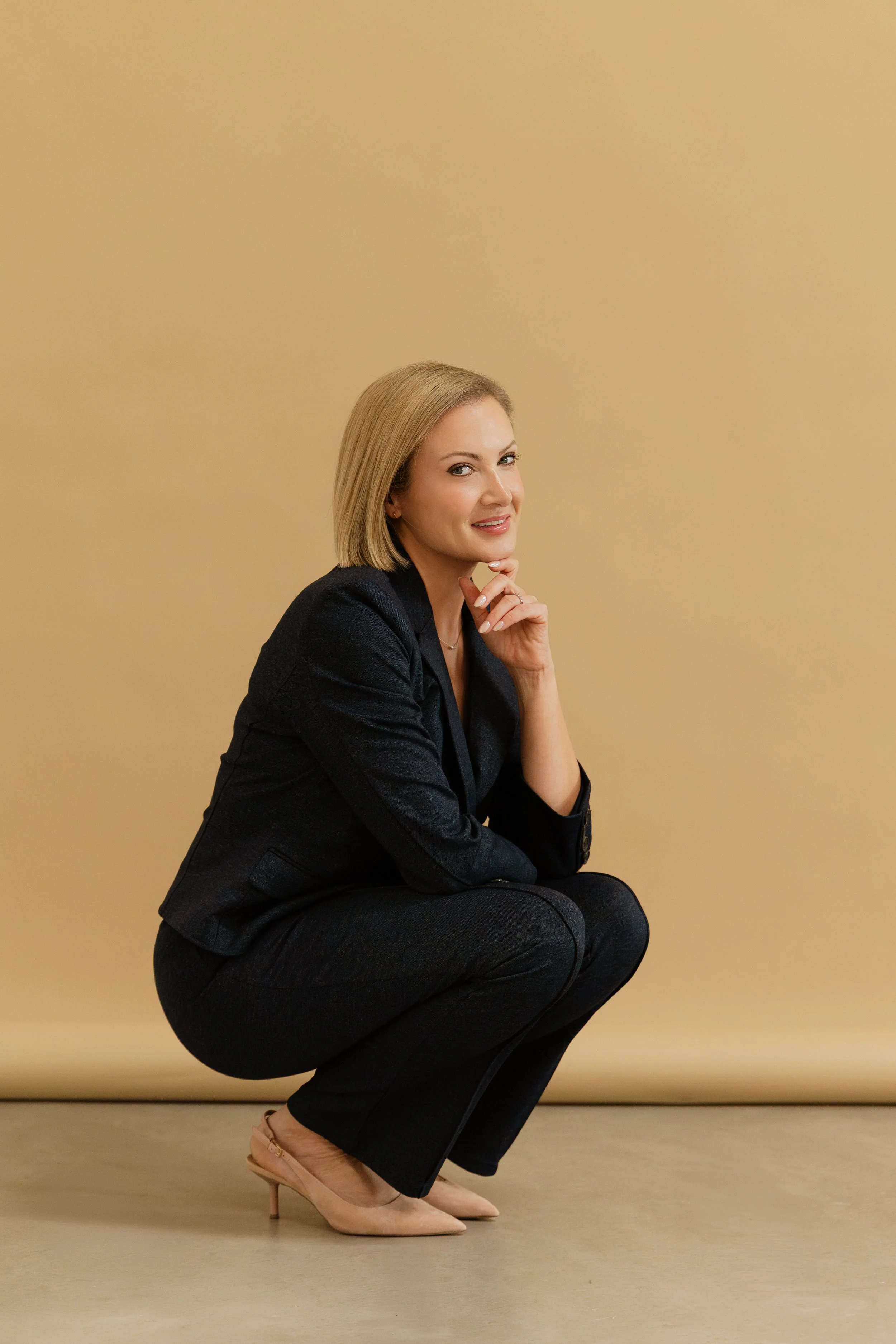 A woman with blonde hair wearing a dark blazer and pants, squatting on the floor with a beige background.