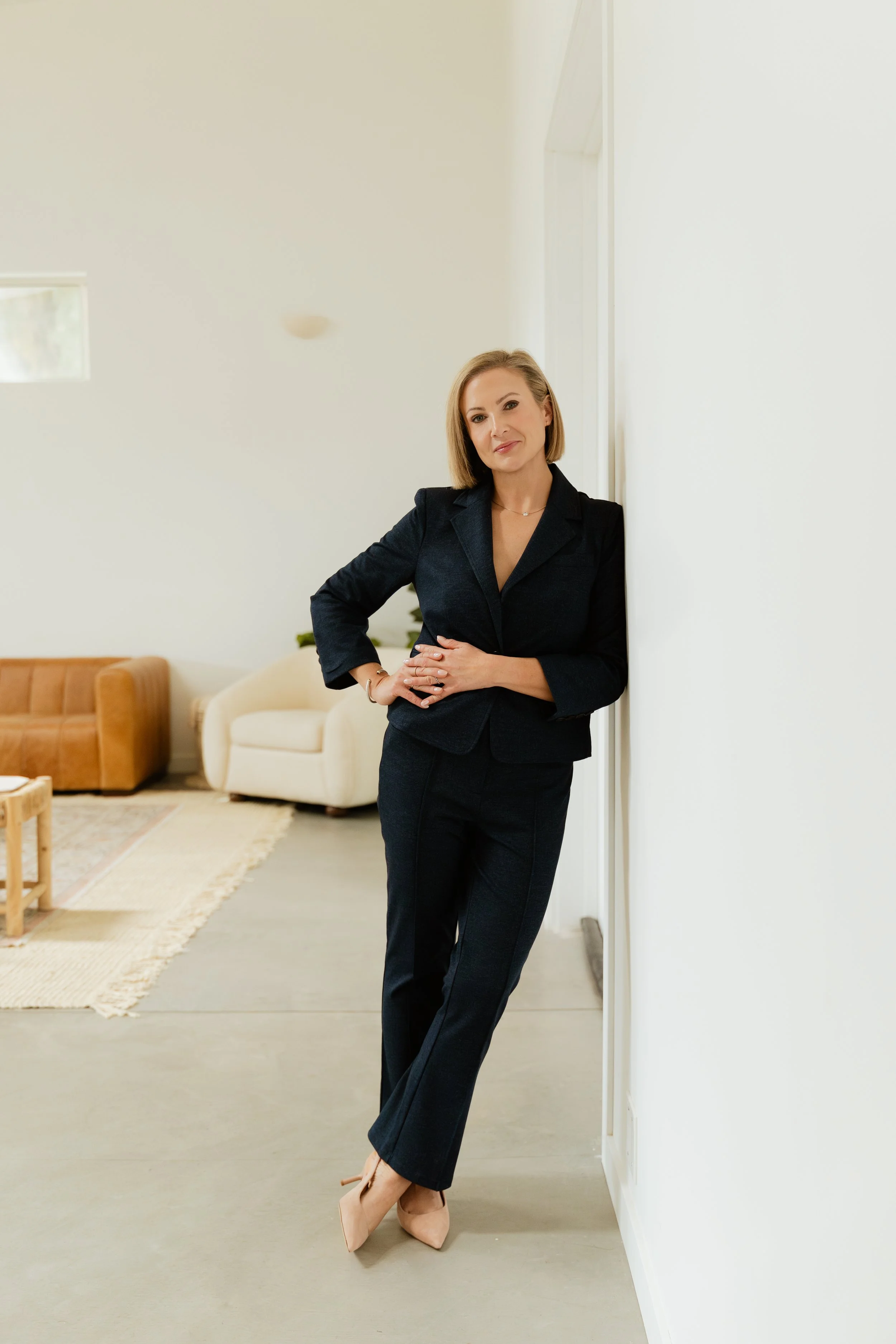 A woman in a dark business suit stands leaning against a white wall in a bright living room with sofas and a rug.
