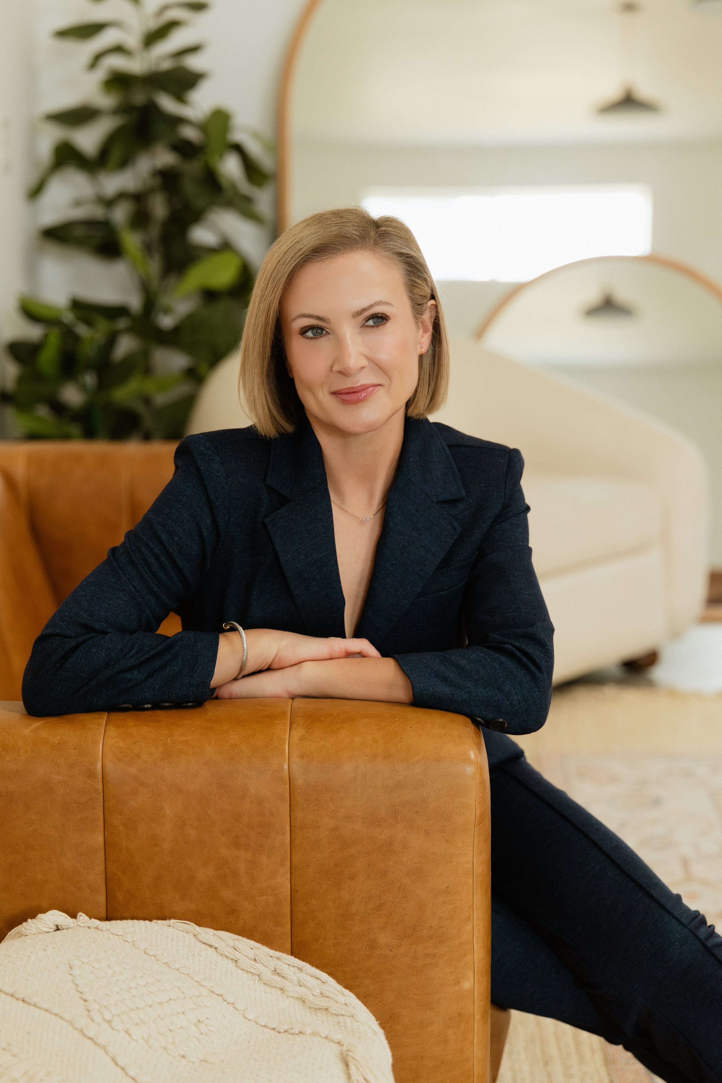 A woman with shoulder-length blonde hair wearing a dark blazer, sitting on a tan leather couch with her arms resting on the back, in a modern living room with a different cream-colored furniture, a large houseplant, and abstract wall art in the background.