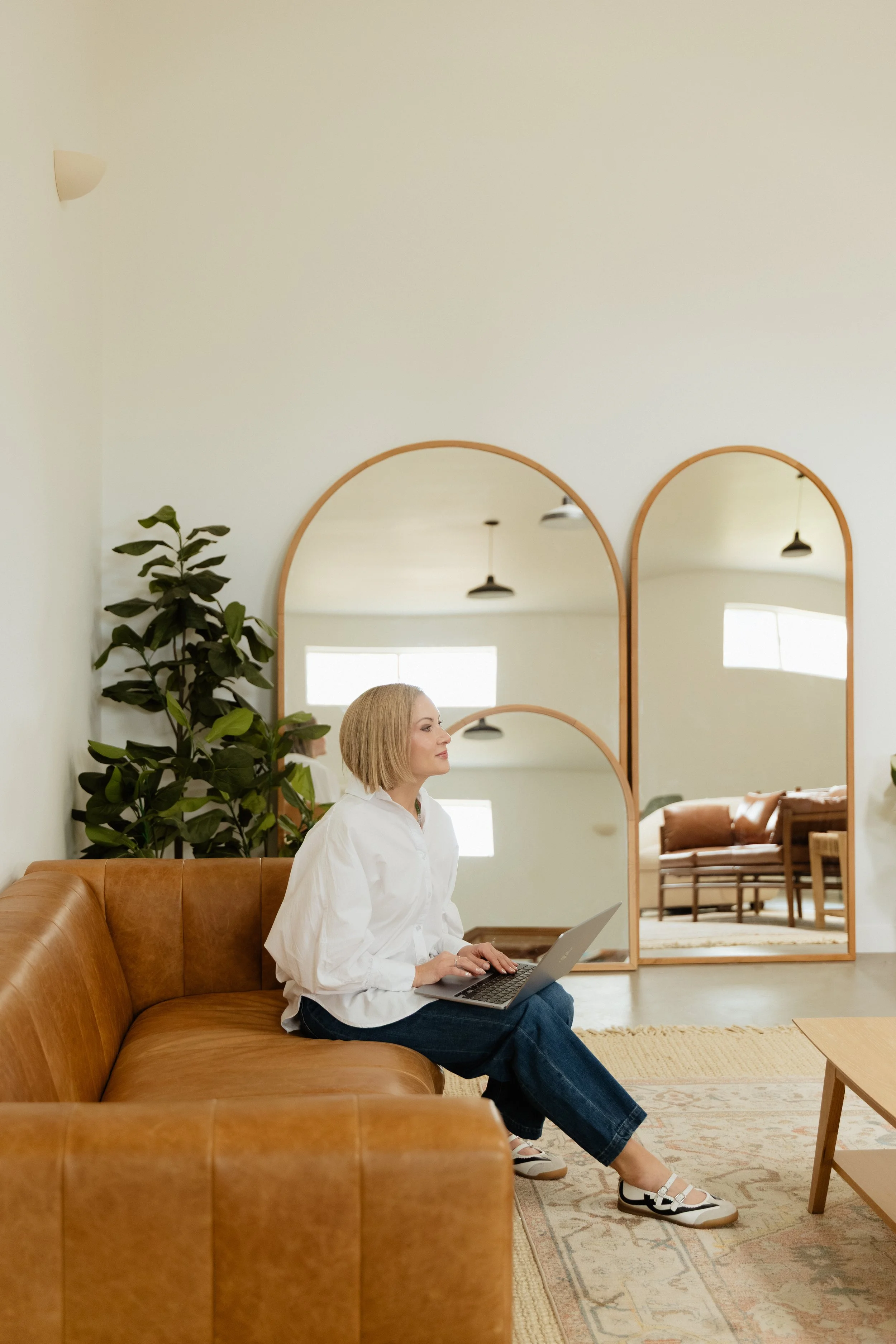 A woman with short blonde hair sitting on a brown leather couch using a laptop, with three large arched mirrors and some green plants in the background, in a bright and modern living room.