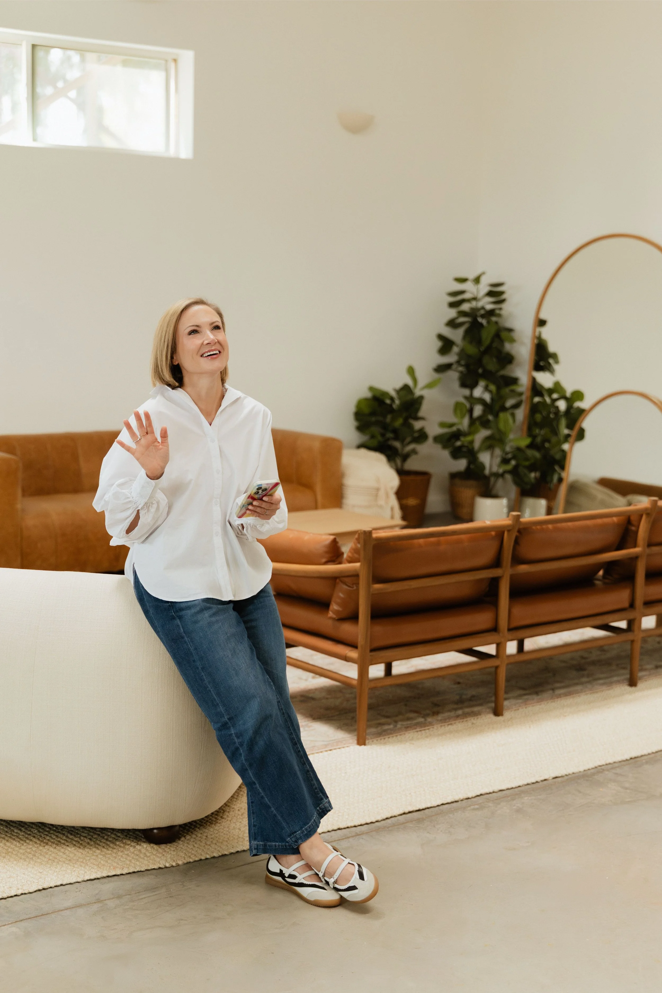 Woman in white shirt and jeans smiling and waving while holding a smartphone in a modern living room.