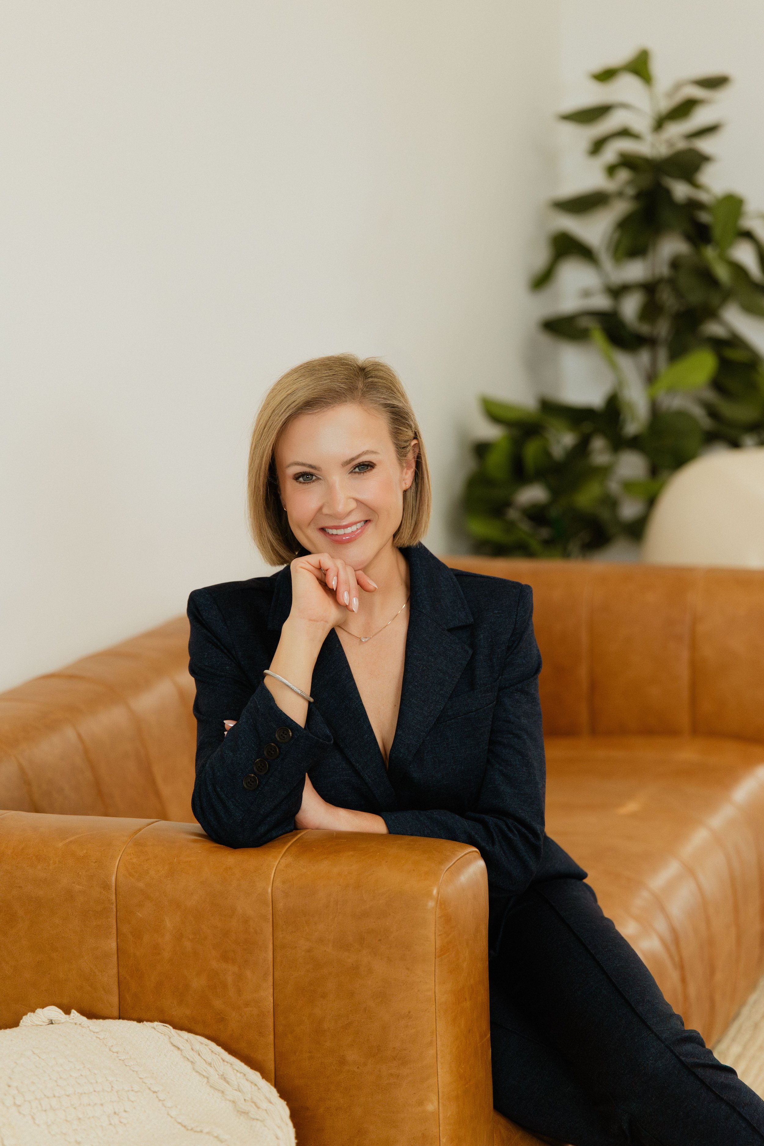 A woman with blonde hair, wearing a dark blazer, sitting on a tan leather sofa, smiling and resting her chin on her hand, with a large leafy plant in the background.