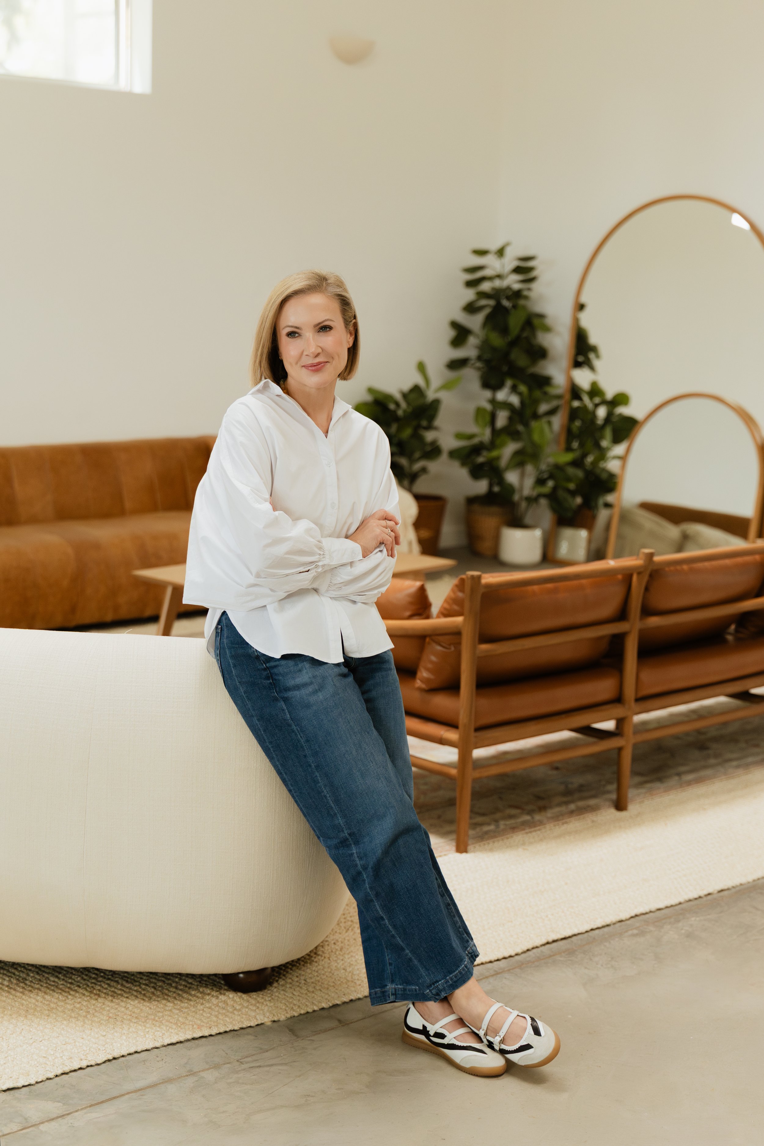 A woman with shoulder-length blond hair wearing a white blouse and blue jeans, leaning against a cream-colored rounded sofa in a modern living room with a brown sofa, green potted plants, a wooden bench, and large mirrors.