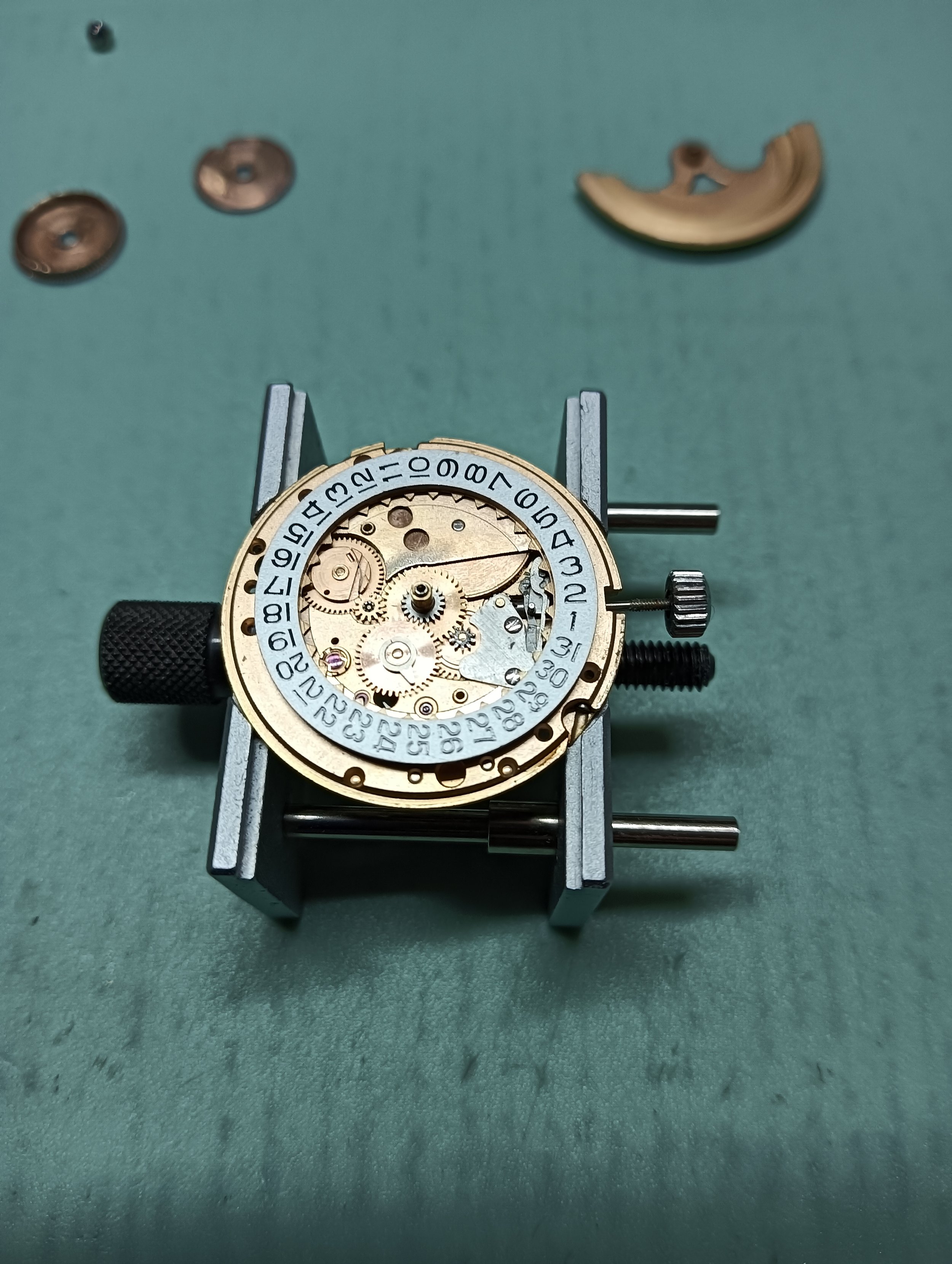 Disassembled watch movement with gears, wheels, and date ring on a green work surface.