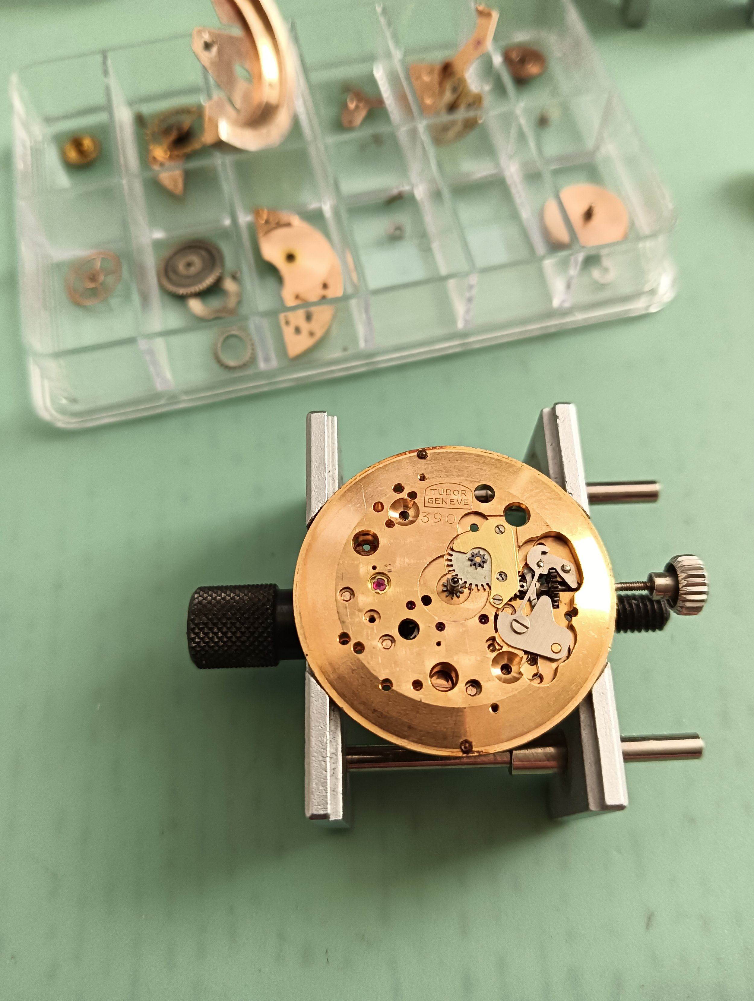 Disassembled watch movement parts on a plastic compartment tray with watchmaking tools in the foreground.