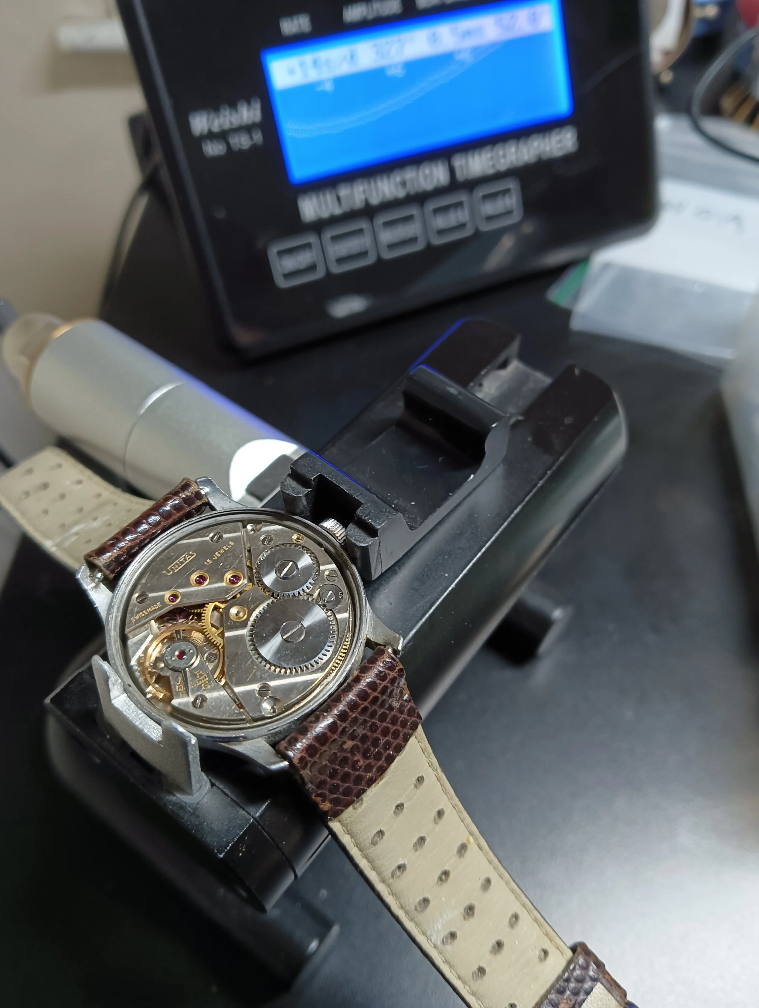 A wristwatch with an exposed mechanical internal mechanism, a leather strap with a textured pattern, positioned on a watchmaker's holder beneath a digital device with a blue display.
