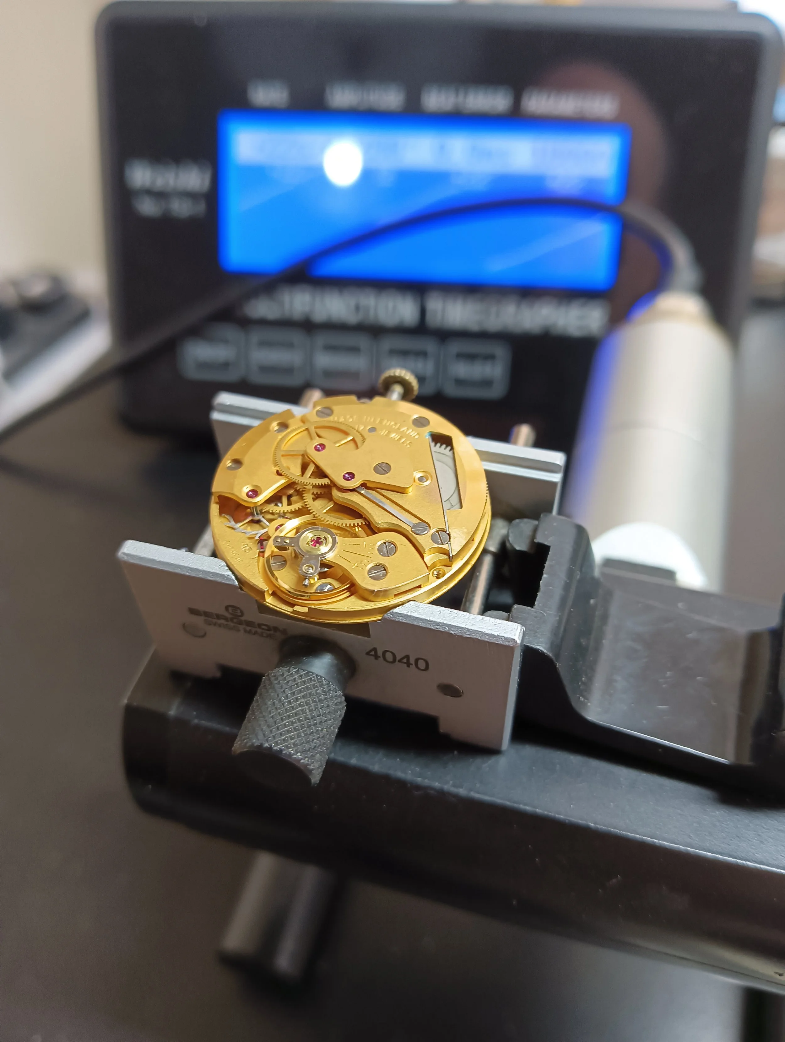 Close-up view of a watch movement held in a vise, with an electronic device displaying a blue screen in the background.