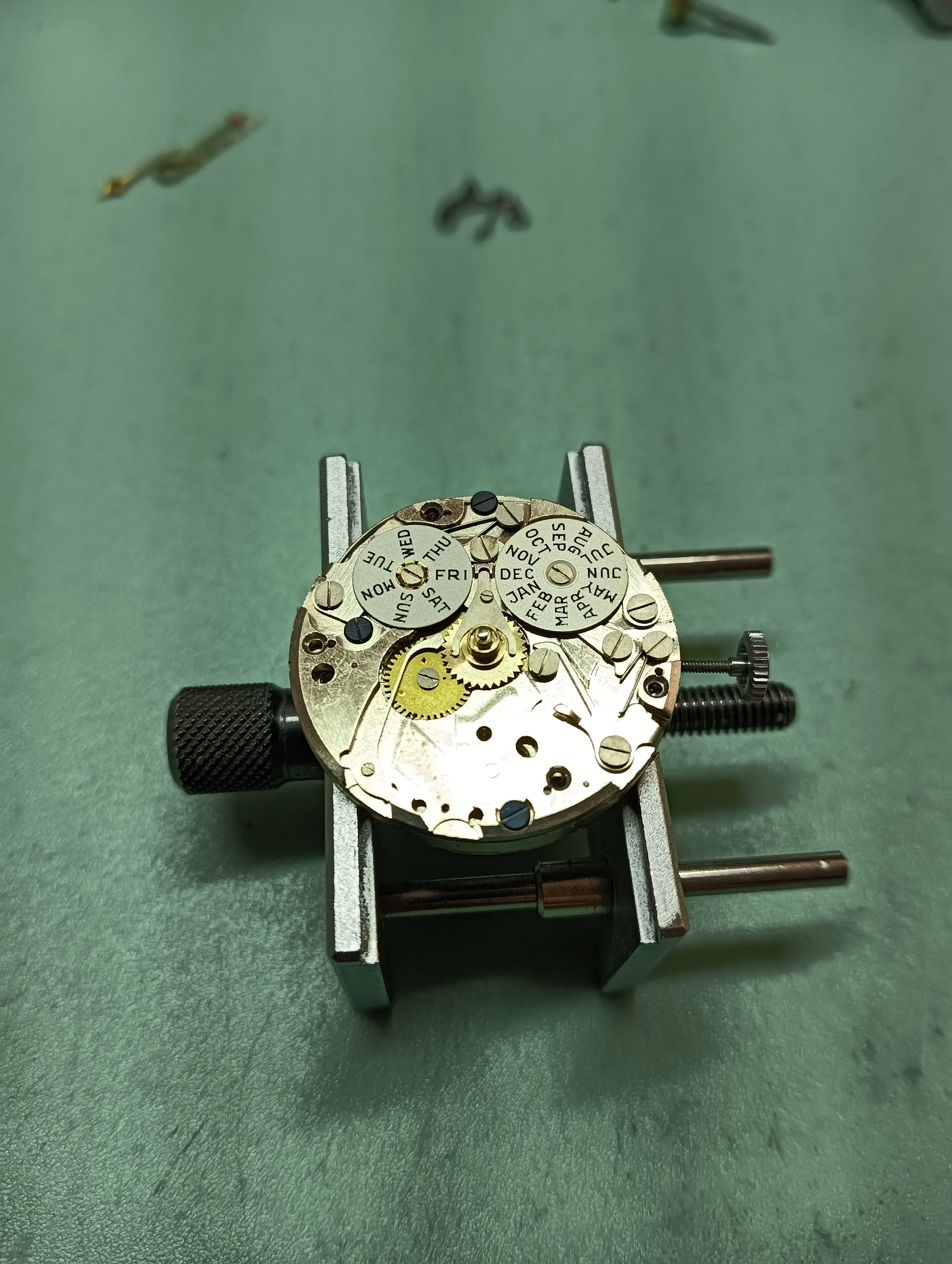 Close-up of a disassembled watch movement with gears and date wheels, placed on a green workbench, held in a watchmaker's holder.